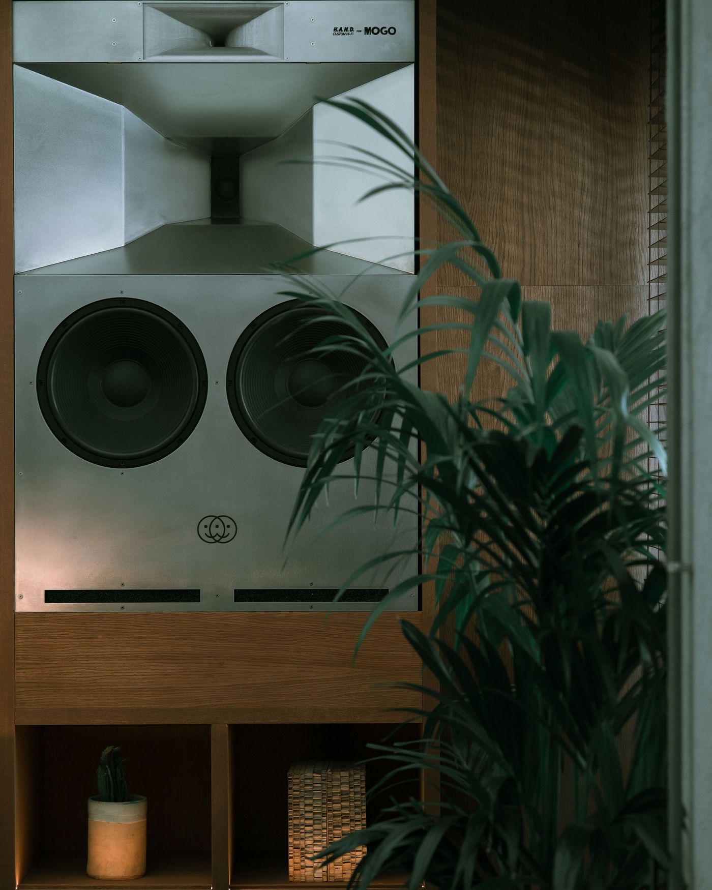 A sleek audio speaker system with horn and dual woofers, surrounded by greenery and warm wood accents, in a cozy setting.
