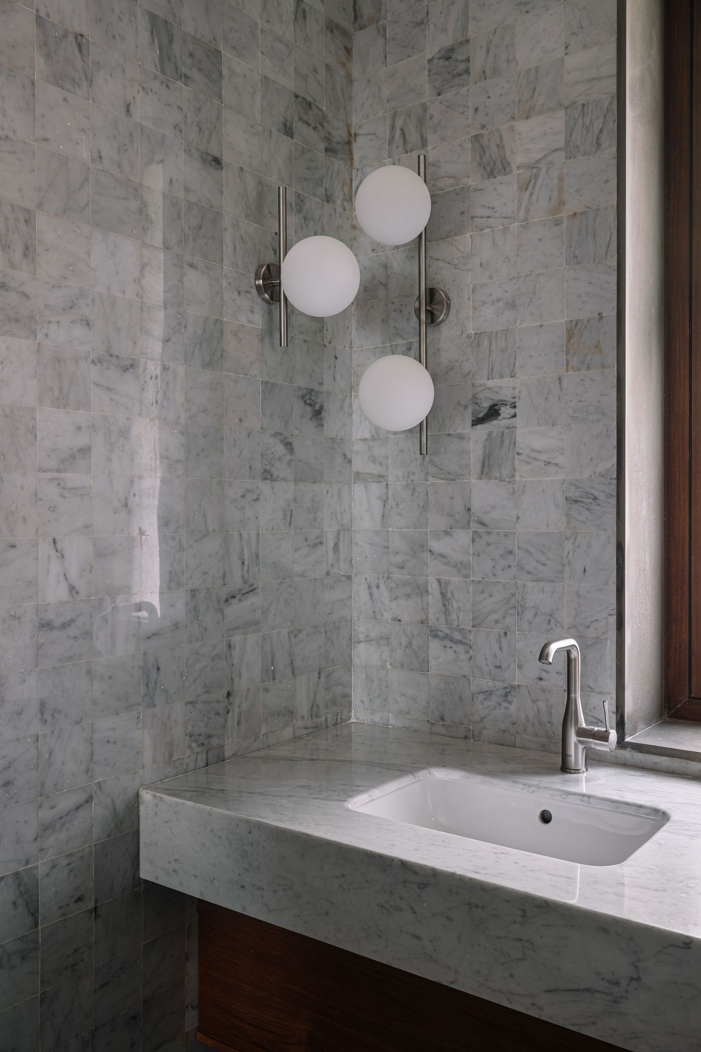 A modern bathroom vanity area features a sleek white rectangular sink integrated into a light-colored countertop. The walls are clad in small, rectangular white marble tiles with subtle grey veining. Two contemporary wall-mounted light fixtures with spherical white diffusers provide illumination, while a wooden window frame on the right adds a touch of warmth and natural light.