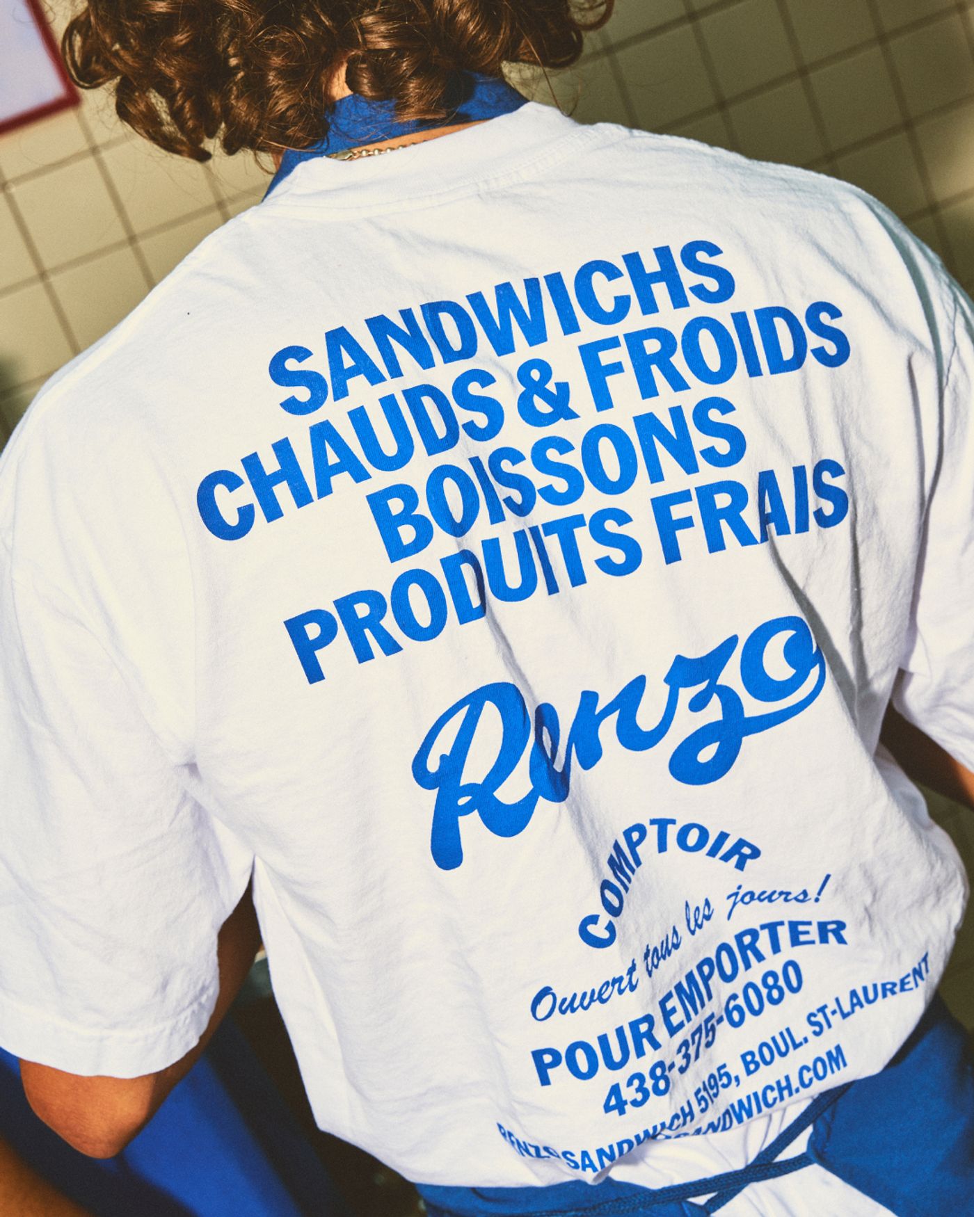 A person's back is shown wearing a white t-shirt with bold blue lettering advertising "SANDWICHS CHAUDS & FROIDS BOISSONS PRODUITS FRAIS." The name "Renzo" is displayed in a distinctive script. Below, smaller text details operating hours and contact information, with a blue apron visible.