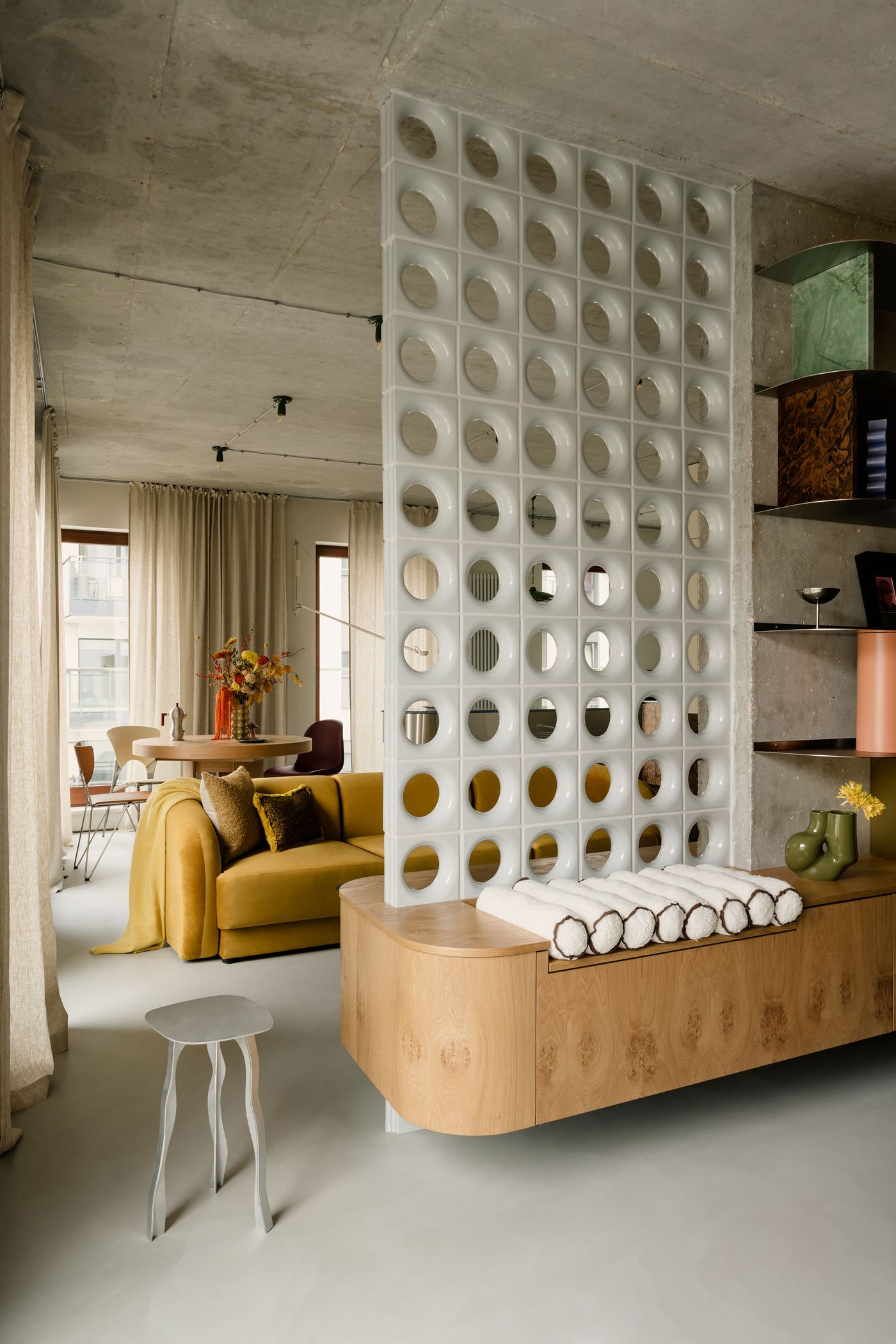 A perforated room divider with circular cutouts adds a playful architectural rhythm to this open-plan space. Blending retro futurism with contemporary softness, the area features a mustard velvet sofa, midcentury-inspired details, and a muted palette grounded by pale wood and concrete textures.