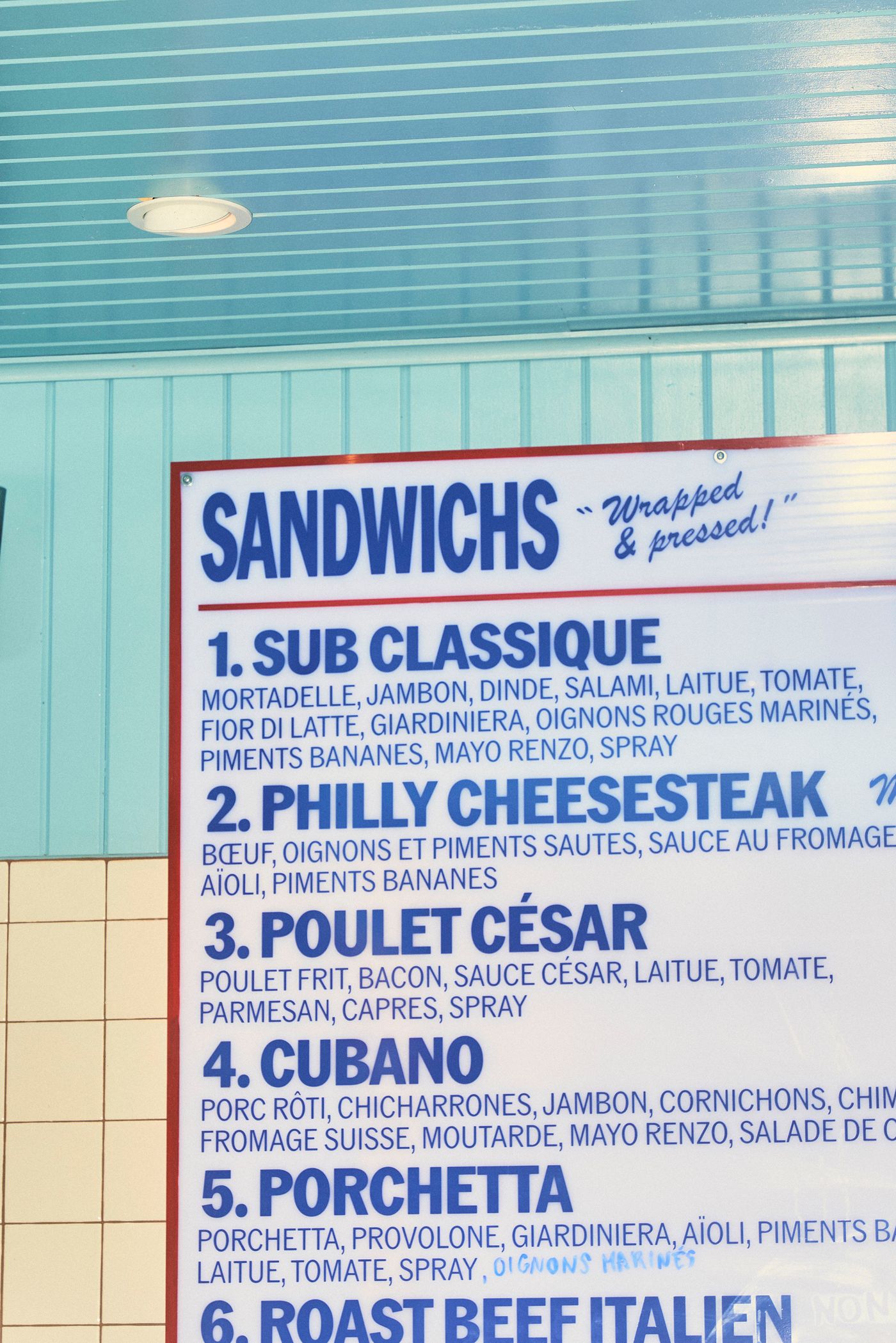 A close-up shot of a menu board inside Renzo Sandwich shop. The menu, predominantly in blue sans-serif font, lists various sandwiches with ingredients. Above the menu, a light blue, slatted ceiling and a white light fixture suggest a retro diner aesthetic.