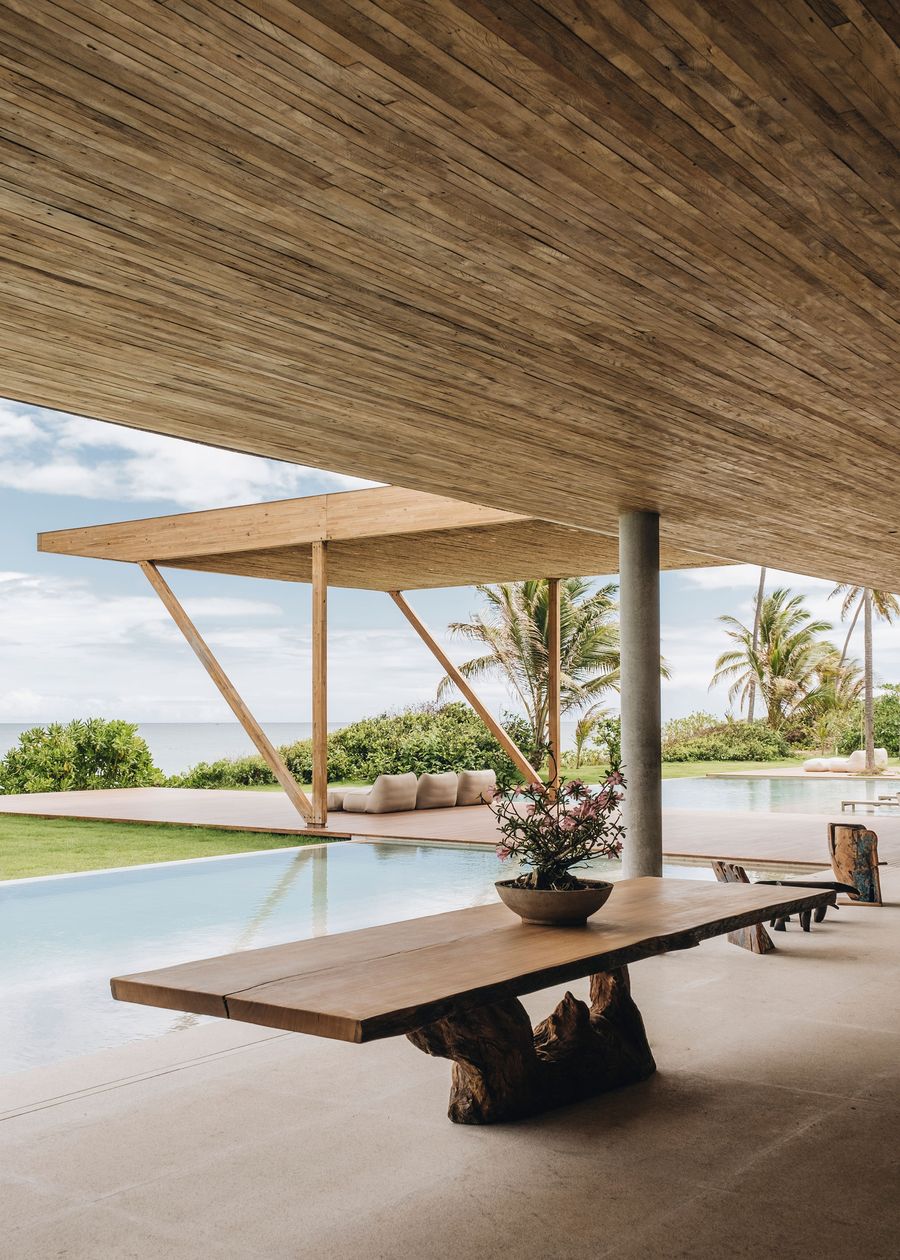 Modern outdoor space featuring a wooden table, pool, and views of tropical greenery and ocean under a spacious wooden ceiling.