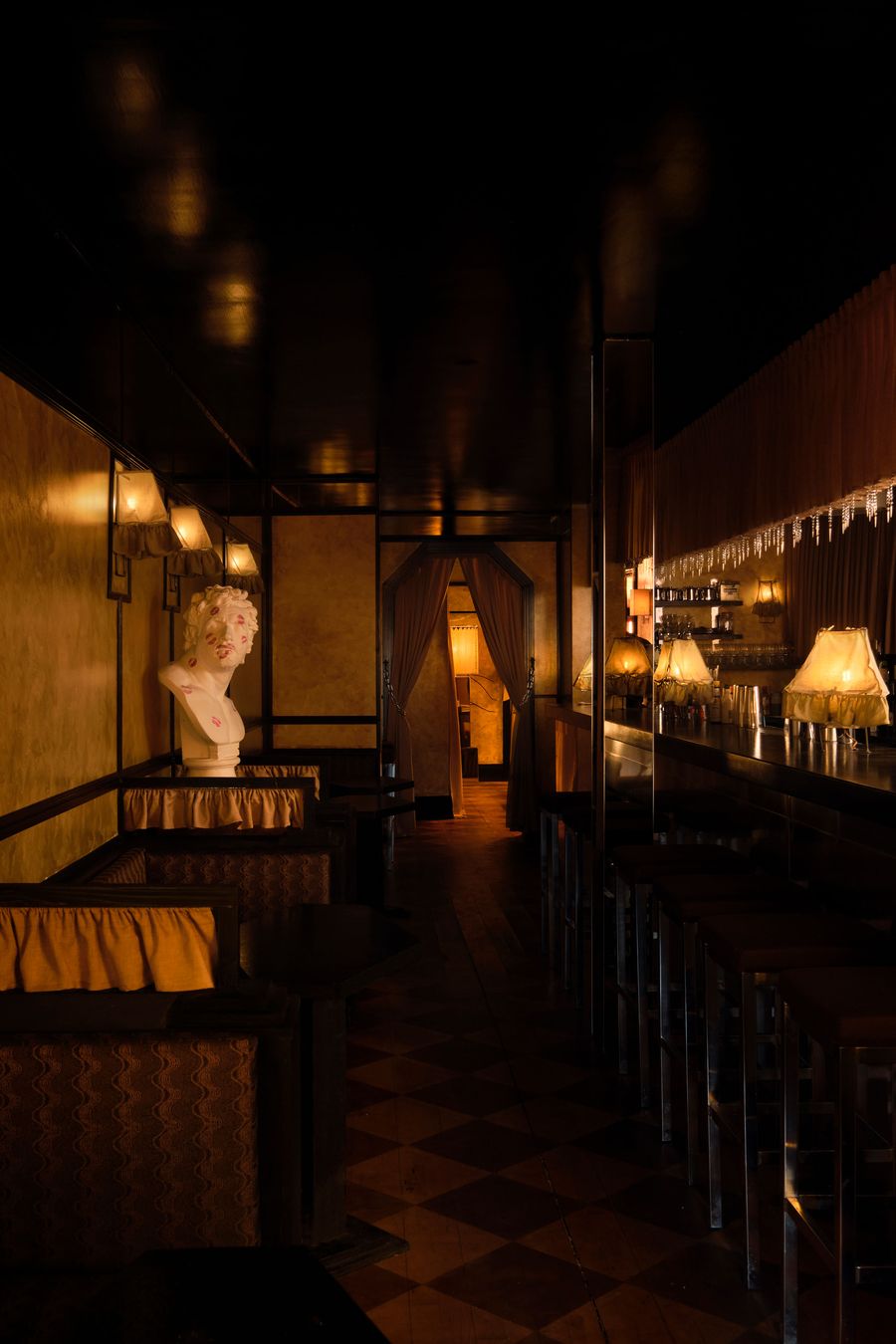 An evocative, dimly lit long shot of The Roses of Elagabalus, showcasing its luxurious vintage-retro aesthetic. On the left, a classical white bust sits atop a draped surface, contrasting with the dark, reflective bar on the right adorned with shimmering icicle-like details and warm, glowing lamps. The checkered floor and rich textures create a decadent, immersive atmosphere.