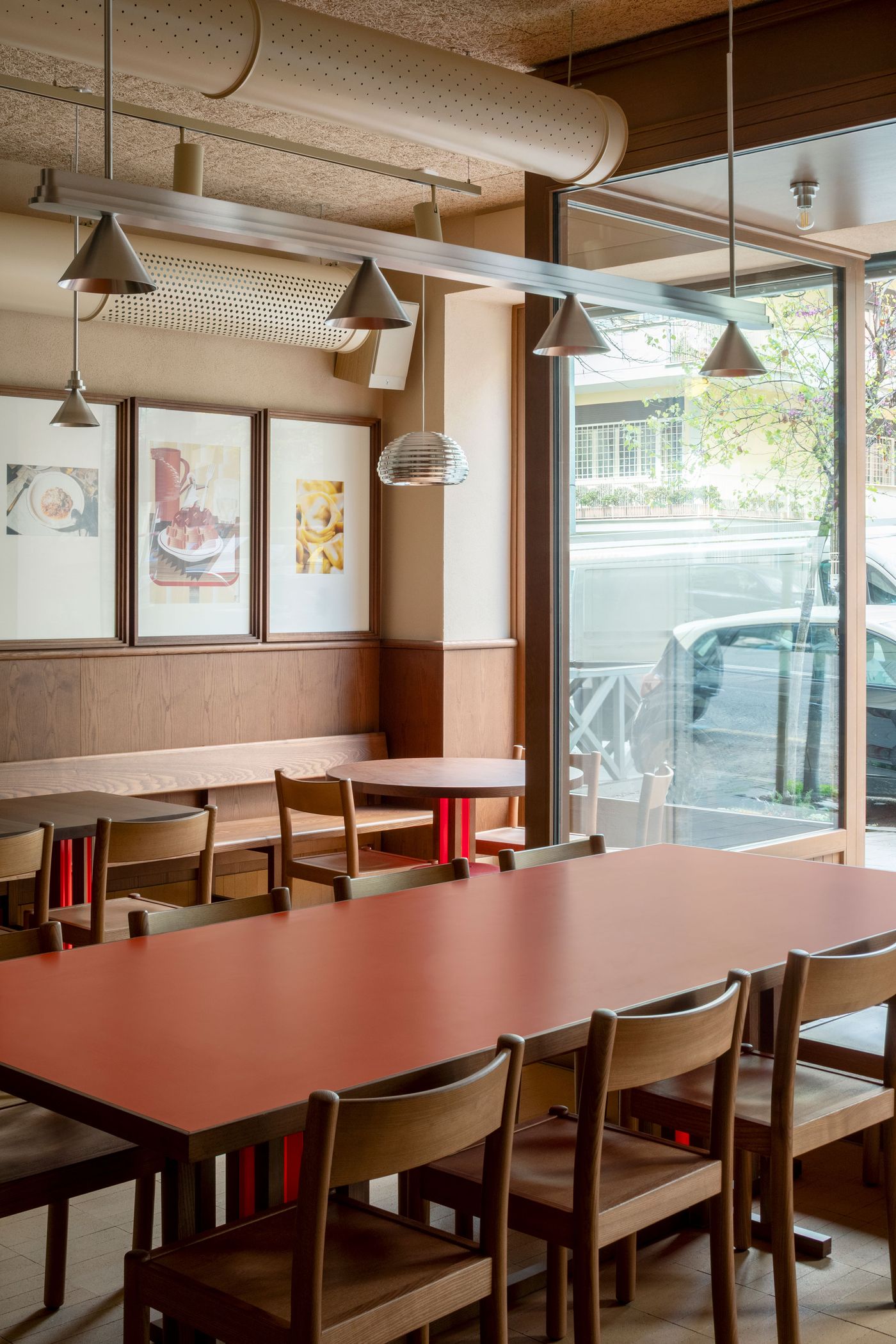 A modern Roman trattoria interior by Naessi Studio, blending warm wood tones with minimalist design. A long orange communal table with subtle red legs occupies the foreground, flanked by light wooden chairs. Industrial pendant lights illuminate the space, with a glimpse of the street outside through large glass panels, enhancing the casual urban atmosphere.