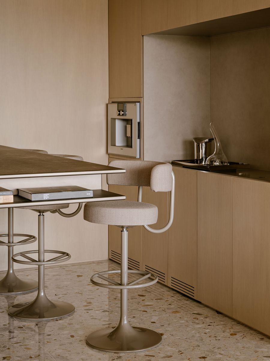 A modern kitchen featuring a minimalist design with a tall, beige stool by a sleek, stone countertop and warm wooden cabinetry. A modern kitchen featuring a minimalist design with a tall, beige stool by a sleek, stone countertop and warm wooden cabinetry.