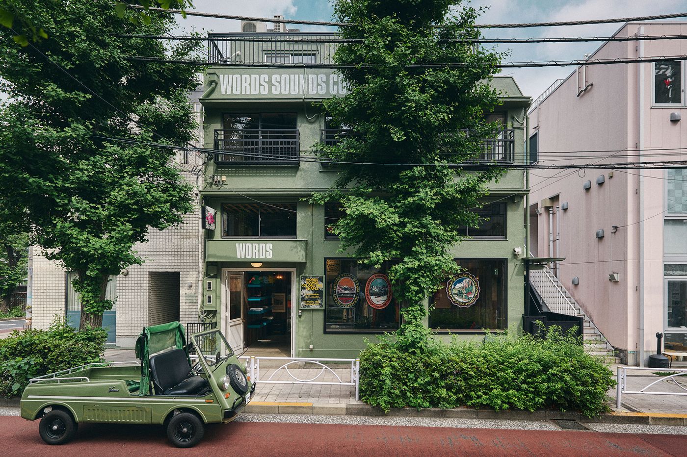 A green storefront labeled "WORDS" with an old-fashioned green vehicle parked outside, surrounded by trees and urban architecture.