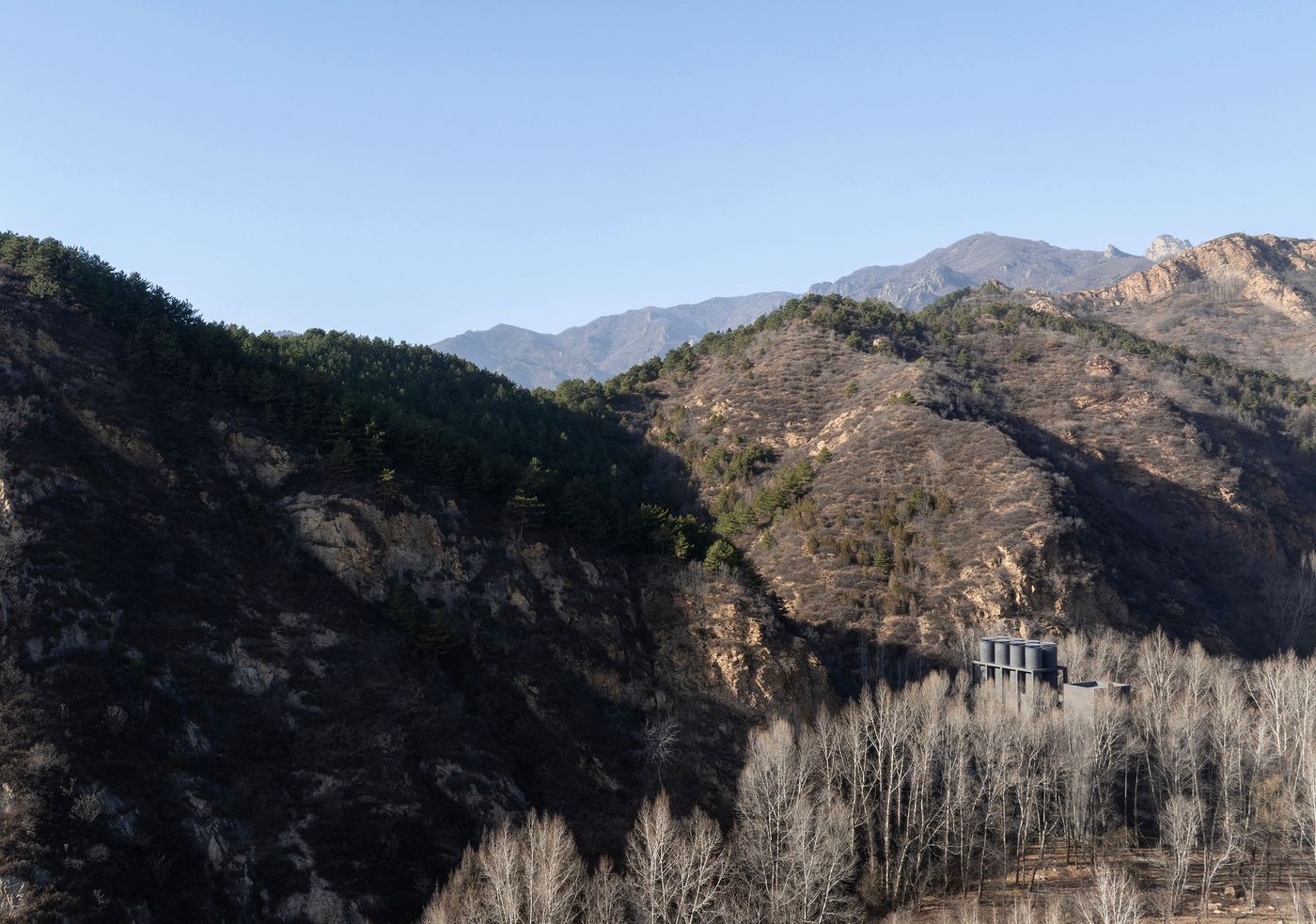 A minimalist gray building nestled among bare trees and green pine-covered hills, with mountains rising in the background under a clear sky. A minimalist gray building nestled among bare trees and green pine-covered hills, with mountains rising in the background under a clear sky.