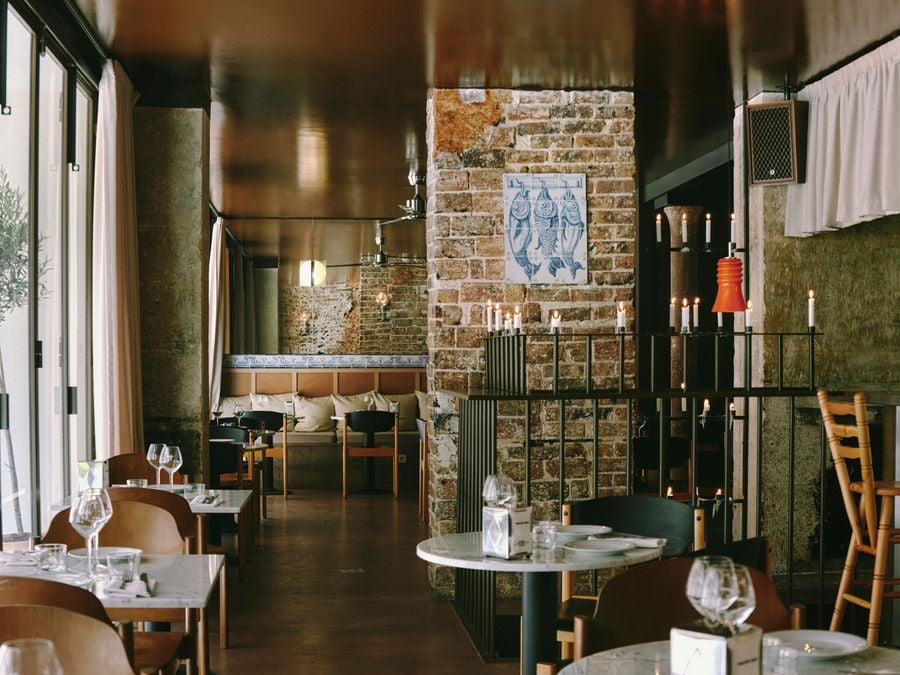 A long view through SaltoBar in Paris, from sunlit window tables towards a cushioned banquette edged in azulejos. A tiled panel of three hanging fish adorns a brick pier. Beside it, a slender metal balustrade bristles with white candles beneath a vivid orange pendant. The mirror-like ceiling doubles the room's warm, amber glow.