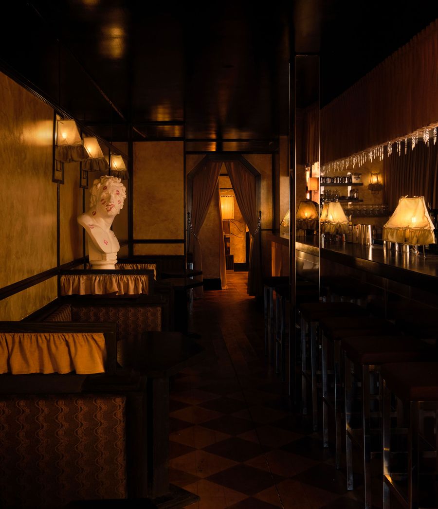 An evocative, dimly lit long shot of The Roses of Elagabalus, showcasing its luxurious vintage-retro aesthetic. On the left, a classical white bust sits atop a draped surface, contrasting with the dark, reflective bar on the right adorned with shimmering icicle-like details and warm, glowing lamps. The checkered floor and rich textures create a decadent, immersive atmosphere.