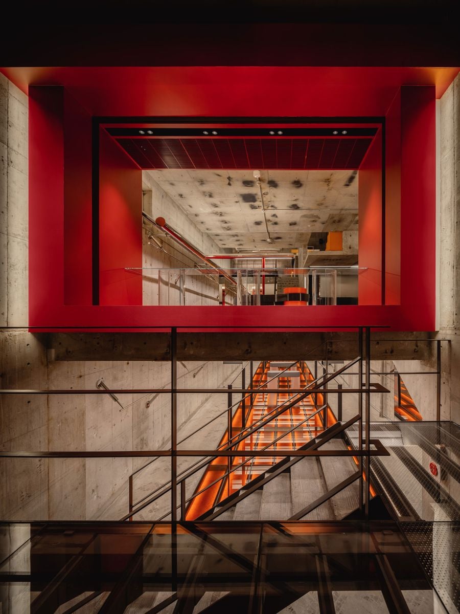 Seen from the street, the red-framed ground floor hovers above the basement, with both levels exposed through the glazed façade. Illuminated stairs descend through the concrete shell, creating a dramatic vertical axis of orange light, while black railings and transparent surfaces emphasise the store’s layered, interconnected spatial structure.