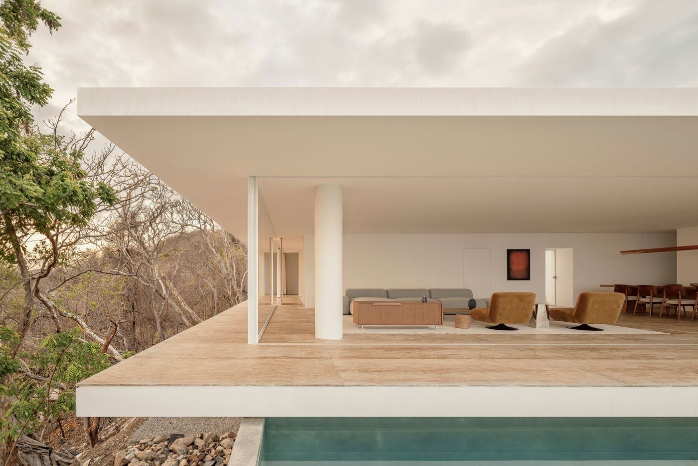 Held between two crisp horizontal slabs, the living level reads like an open stage above the pool, punctuated by a single white column. A grey modular sofa, walnut sideboard and caramel-toned velvet chairs sit before a rust-hued colour-field painting, while a glazed corridor recedes along the façade into the trees. Held between two crisp horizontal slabs, the living level reads like an open stage above the pool, punctuated by a single white column. A grey modular sofa, walnut sideboard and caramel-toned velvet chairs sit before a rust-hued colour-field painting, while a glazed corridor recedes along the façade into the trees.