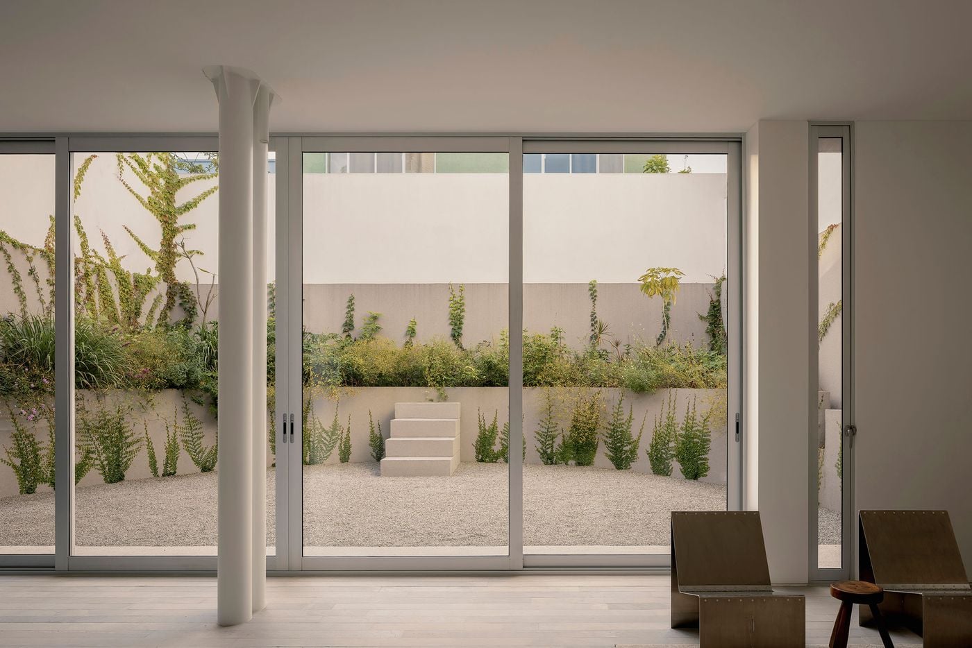 The ground-floor living area opens almost completely onto Casa Tlaloc’s rear courtyard through floor-to-ceiling sliding glass doors. Pale stone flooring and white walls form a restrained interior foreground, while gravel, tiered planting and climbing vegetation soften the high perimeter walls and lend the courtyard a secluded, contemplative atmosphere. The ground-floor living area opens almost completely onto Casa Tlaloc’s rear courtyard through floor-to-ceiling sliding glass doors. Pale stone flooring and white walls form a restrained interior foreground, while gravel, tiered planting and climbing vegetation soften the high perimeter walls and lend the courtyard a secluded, contemplative atmosphere.
