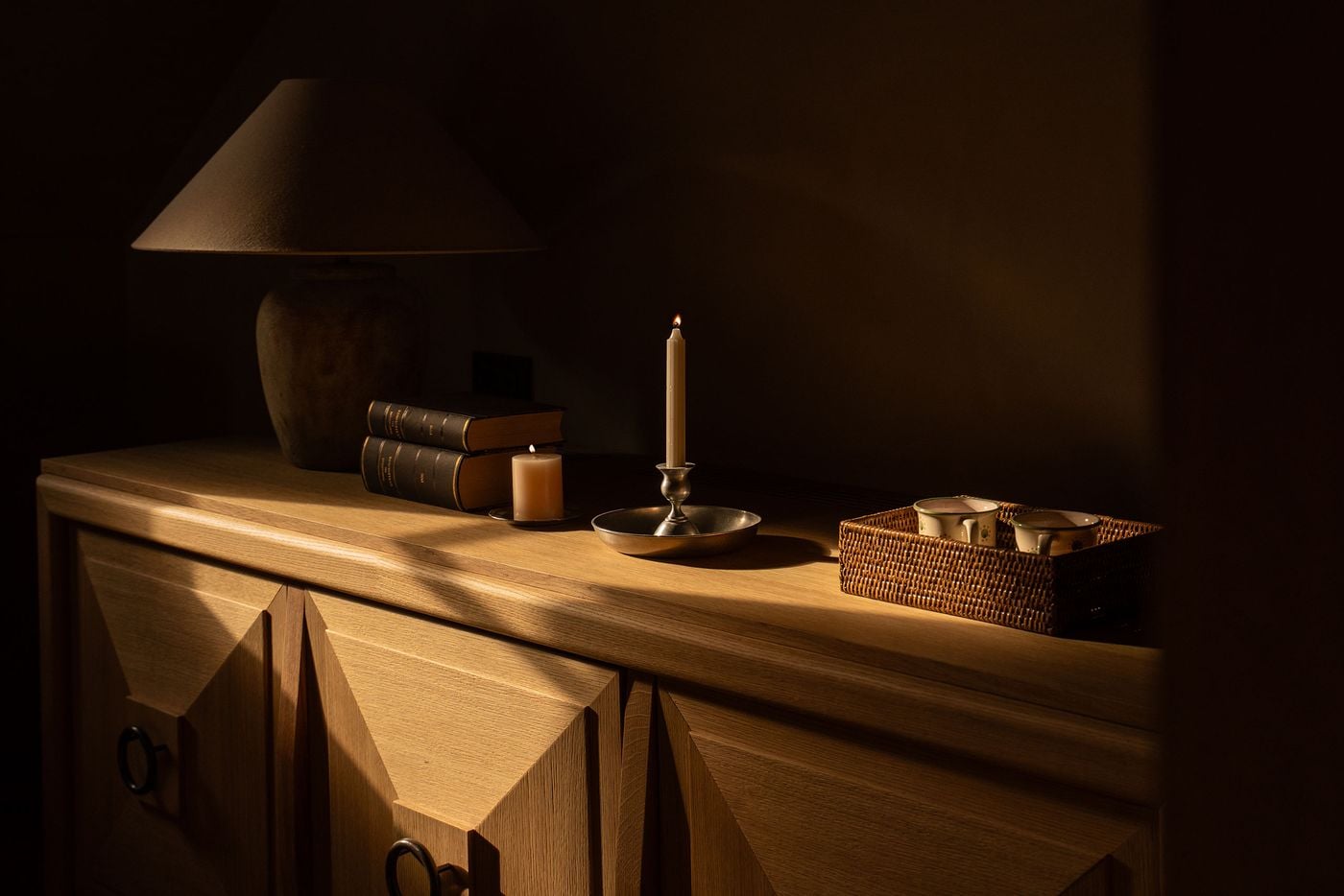 Raking light catches an oak sideboard at Castel Badia, its doors carved into faceted diamond-point panels with dark iron ring pulls. On top sit a stoneware lamp with a linen shade, leather-bound books, a pewter chamberstick, a pillar candle and a rattan tray holding two speckled ceramic mugs. Raking light catches an oak sideboard at Castel Badia, its doors carved into faceted diamond-point panels with dark iron ring pulls. On top sit a stoneware lamp with a linen shade, leather-bound books, a pewter chamberstick, a pillar candle and a rattan tray holding two speckled ceramic mugs.