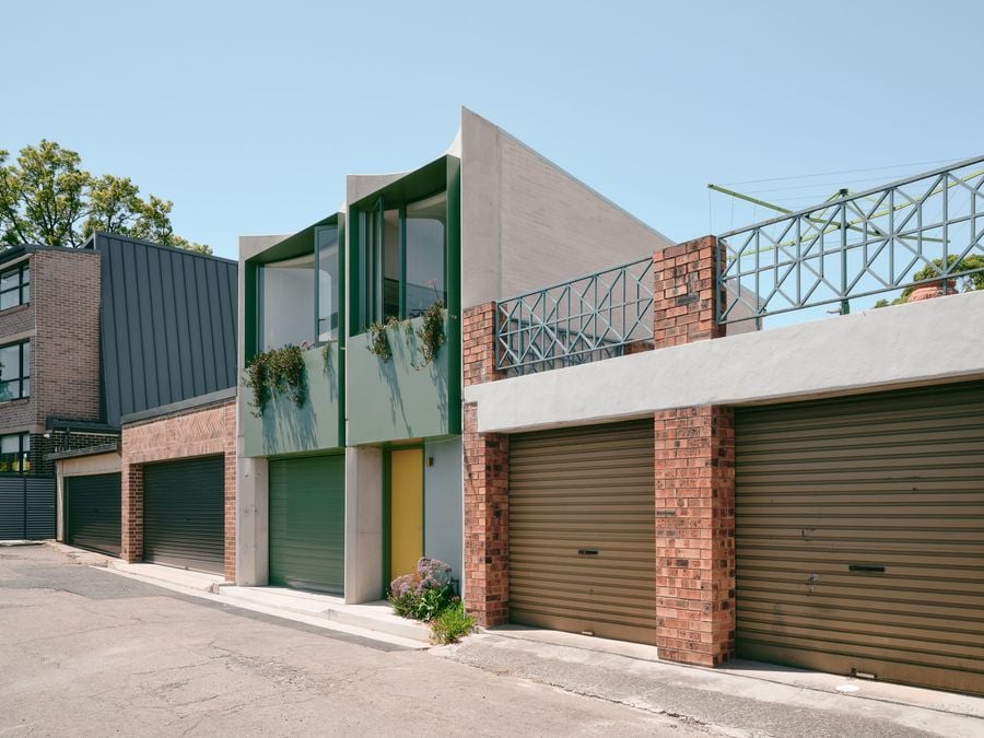 An oblique laneway view places Denj Lane within its garage-lined suburban context, highlighting how the two-storey addition departs from its subdued neighbours. Curved concrete profiles, deep green window surrounds, trailing planting and a yellow entrance bring a distinctly domestic, animated presence to the otherwise functional rear streetscape.