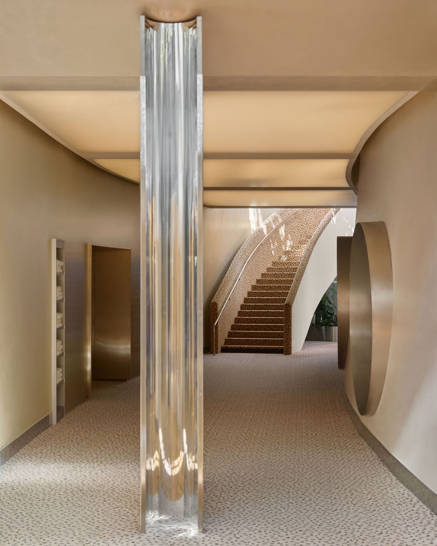 A stainless steel column anchors one of the Ken Club’s principal circulation spaces, catching and refracting daylight against warm neutral walls. In the background, the upholstered staircase rises beneath a softly illuminated ceiling, while a circular wall opening continues the renovation’s recurring language of rounded geometry. A stainless steel column anchors one of the Ken Club’s principal circulation spaces, catching and refracting daylight against warm neutral walls. In the background, the upholstered staircase rises beneath a softly illuminated ceiling, while a circular wall opening continues the renovation’s recurring language of rounded geometry.