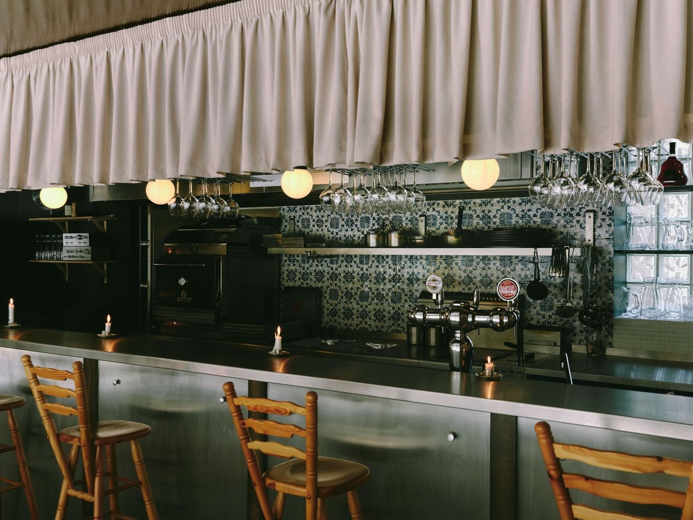 The open kitchen and bar at SaltoBar in Paris, where a gathered cream fabric valance softens a line of opal globe lights and suspended wine glasses. Below, a backsplash of intricately patterned blue-and-white azulejos meets stainless-steel counters, polished beer taps and a charcoal oven. Pine ladder-back stools line the brushed-steel bar. The open kitchen and bar at SaltoBar in Paris, where a gathered cream fabric valance softens a line of opal globe lights and suspended wine glasses. Below, a backsplash of intricately patterned blue-and-white azulejos meets stainless-steel counters, polished beer taps and a charcoal oven. Pine ladder-back stools line the brushed-steel bar.