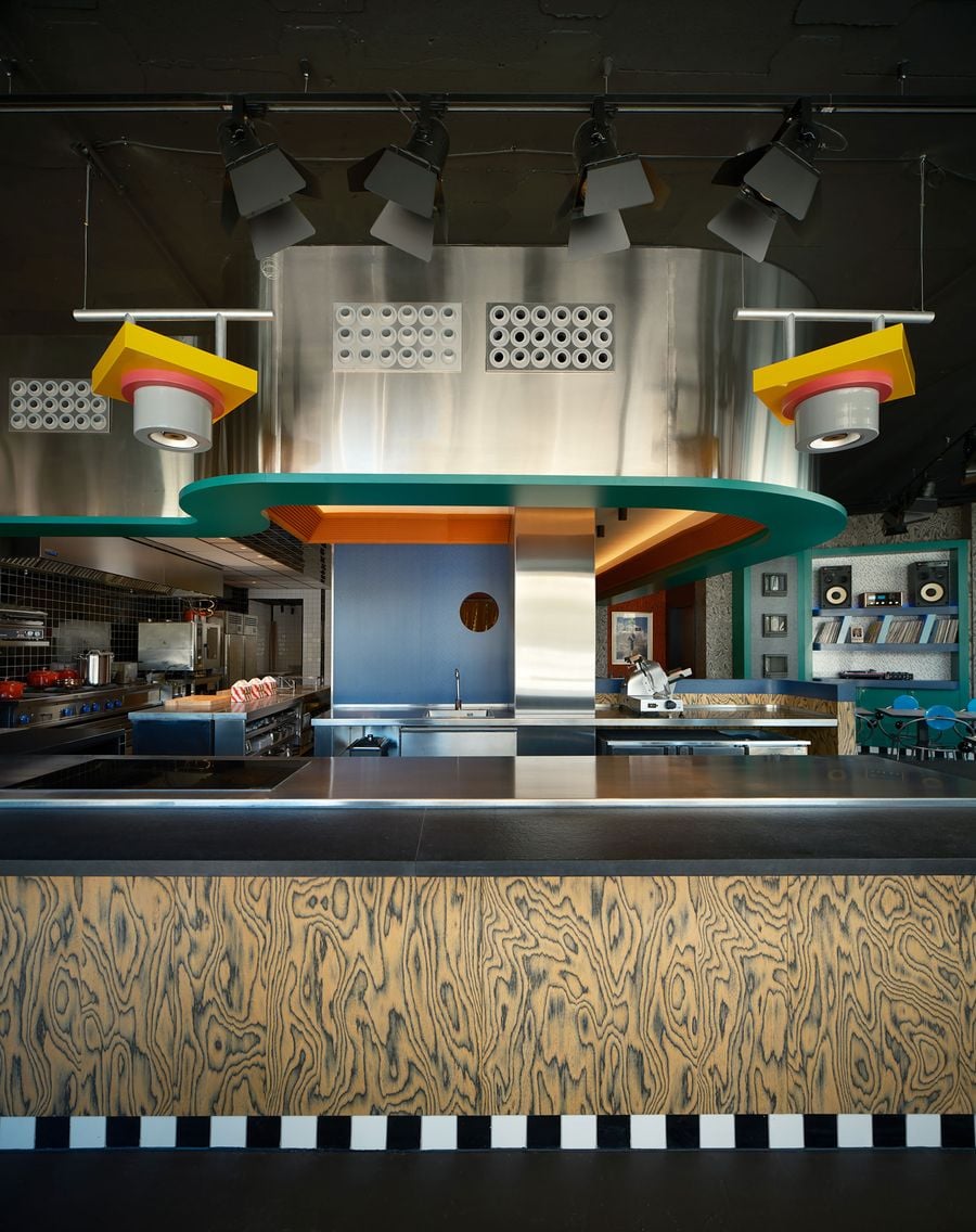 A frontal view of the open kitchen shows an ochre wood-grain counter on a checkerboard skirting, beneath a stainless-steel hood punctuated by perforated vent panels. A teal overhang snakes across the composition between yellow pendant lights. A cobalt wall with a porthole window separates the black-tiled kitchen from the record wall beyond. A frontal view of the open kitchen shows an ochre wood-grain counter on a checkerboard skirting, beneath a stainless-steel hood punctuated by perforated vent panels. A teal overhang snakes across the composition between yellow pendant lights. A cobalt wall with a porthole window separates the black-tiled kitchen from the record wall beyond.