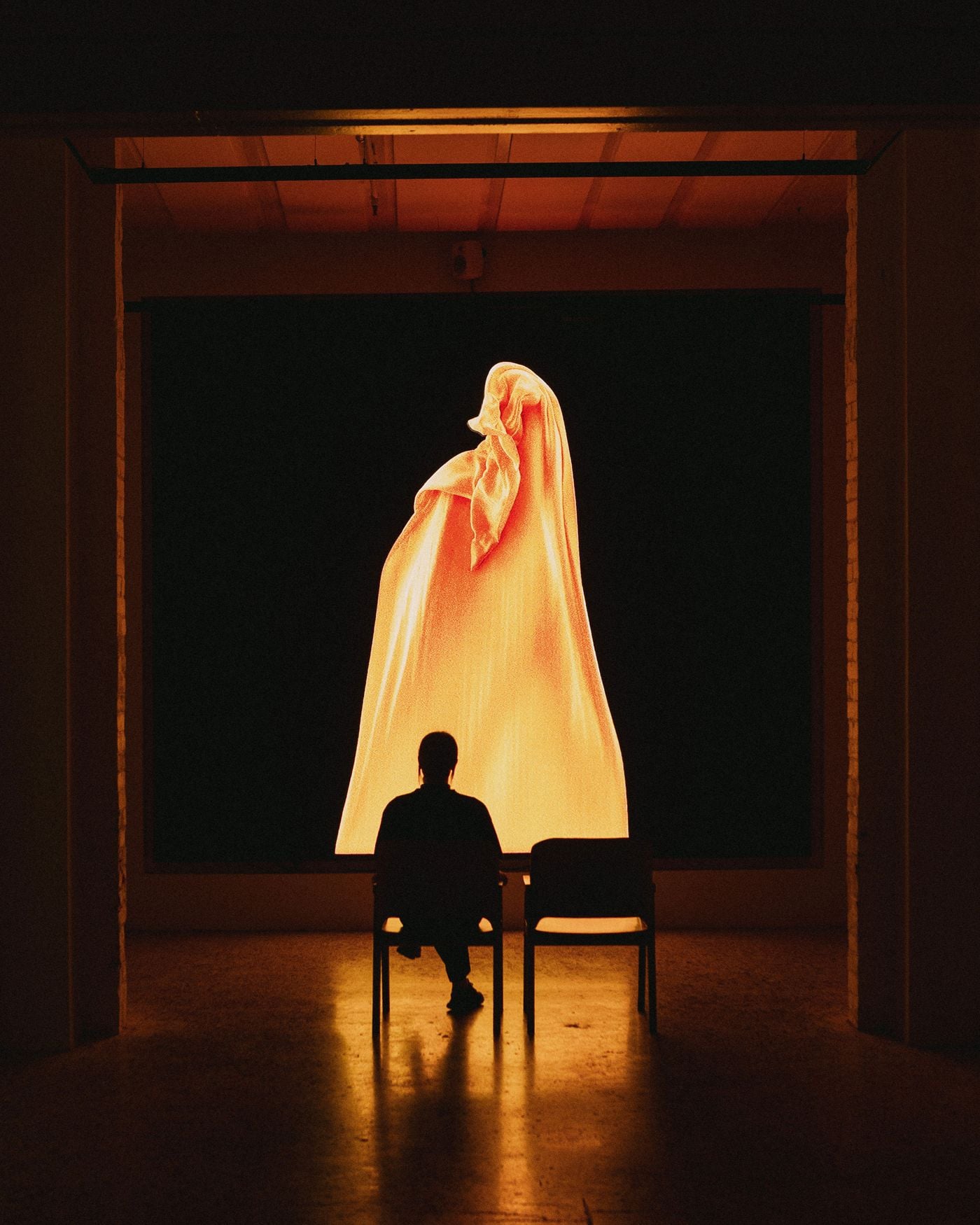 A lone visitor silhouetted on a chair before a large-scale video installation in a darkened gallery, a glowing orange draped form filling the black screen and casting warm reflections across the polished floor and timber ceiling.