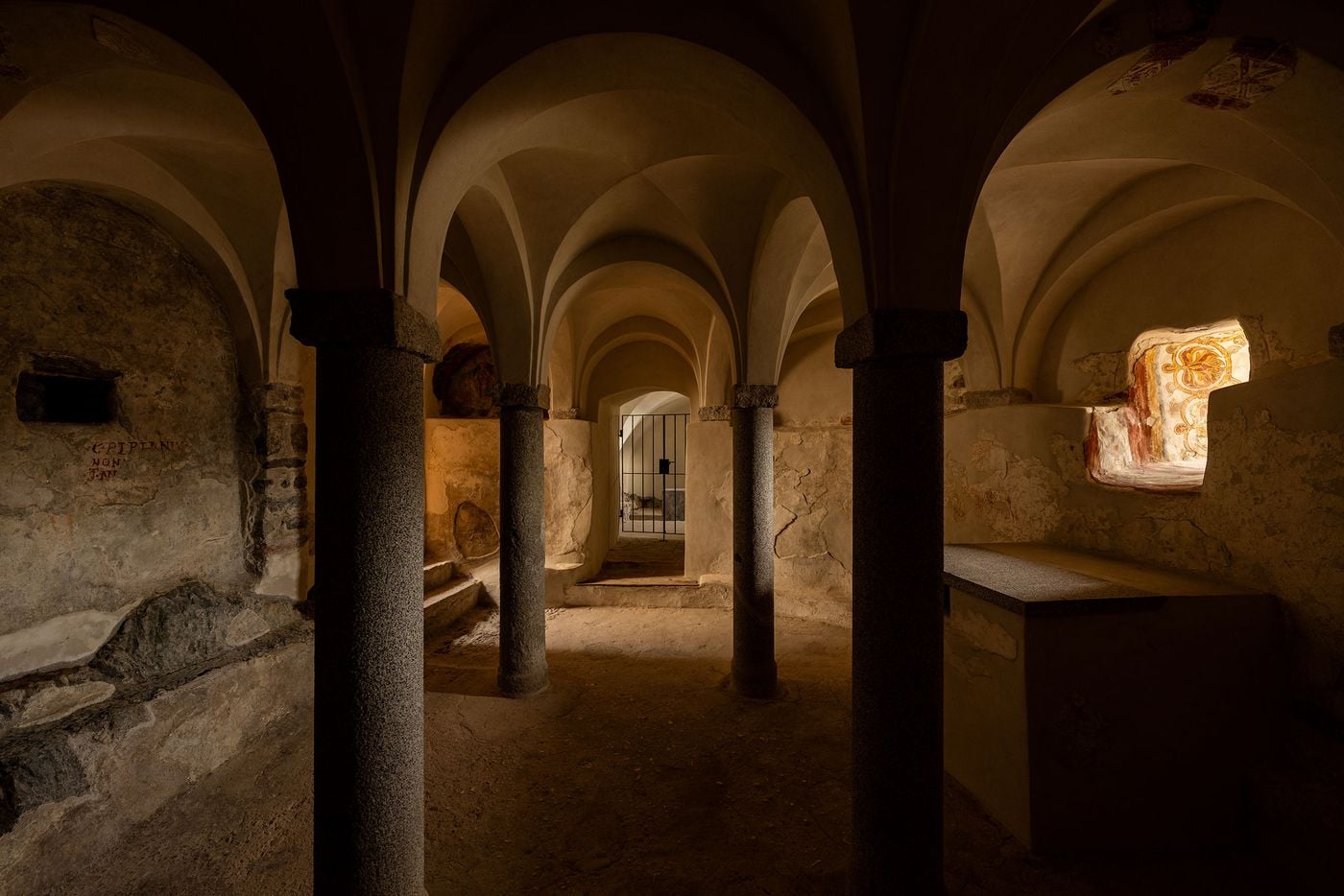 Castel Badia's medieval crypt unfolds as a forest of groin vaults carried on squat granite columns with blocky capitals. Patches of surviving fresco, a scrolling ochre and red ornament and a faded painted inscription mark the time-worn walls, while an iron gate at the far end frames a glimpse of the next chamber. Castel Badia's medieval crypt unfolds as a forest of groin vaults carried on squat granite columns with blocky capitals. Patches of surviving fresco, a scrolling ochre and red ornament and a faded painted inscription mark the time-worn walls, while an iron gate at the far end frames a glimpse of the next chamber.