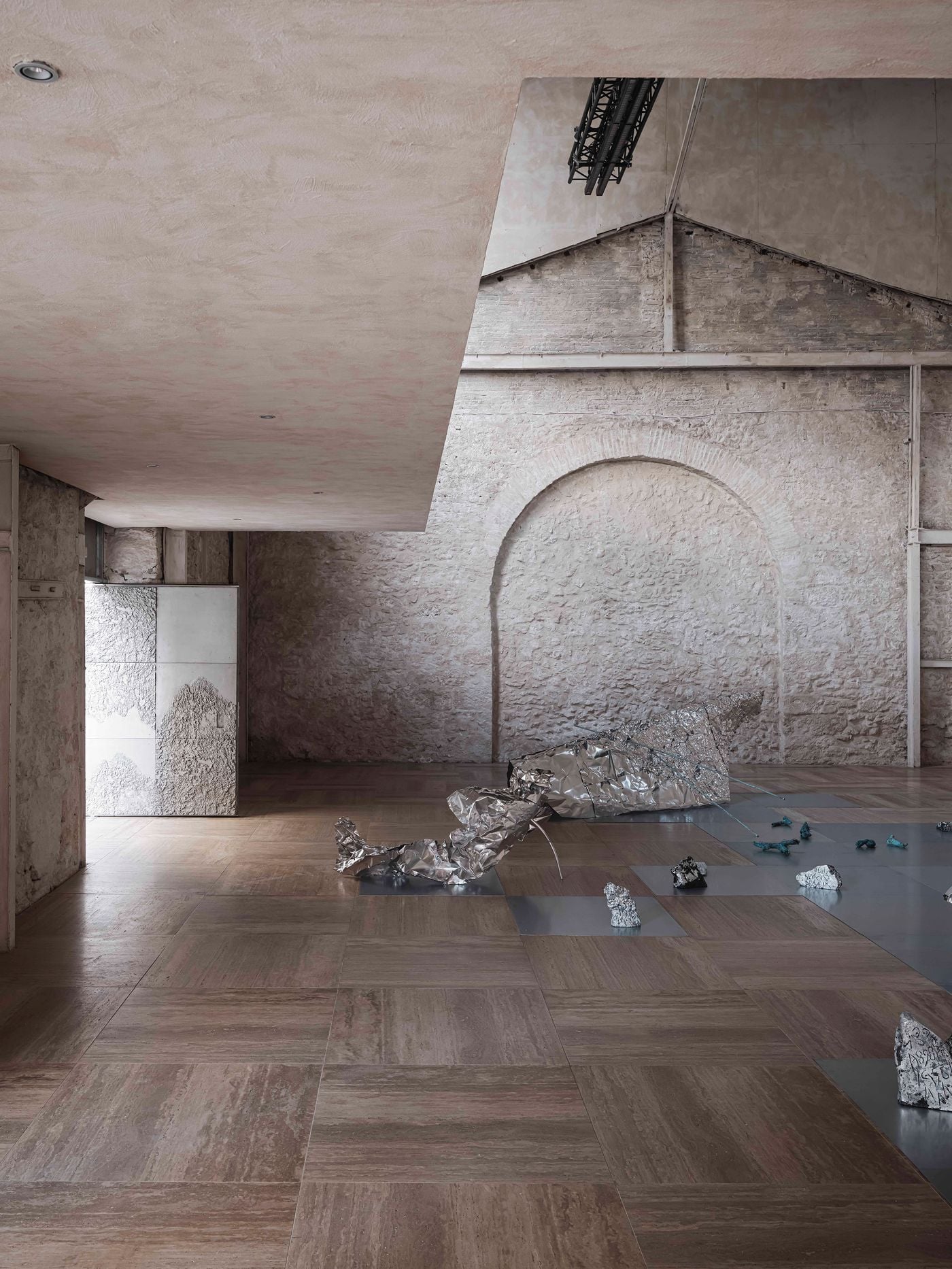 Metallic sculptures from “Casting Water” spread across the travertine floor of Castor Place’s main exhibition hall. Their crumpled, reflective surfaces contrast with the weathered masonry and softly plastered interventions, while the preserved outline of an earlier arched opening anchors the composition in the building’s industrial past. Metallic sculptures from “Casting Water” spread across the travertine floor of Castor Place’s main exhibition hall. Their crumpled, reflective surfaces contrast with the weathered masonry and softly plastered interventions, while the preserved outline of an earlier arched opening anchors the composition in the building’s industrial past.