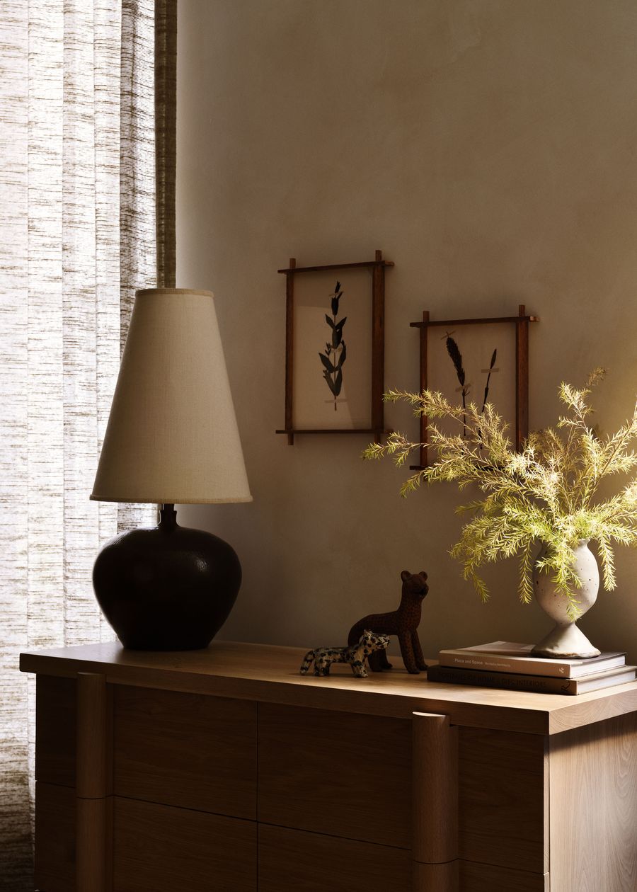 Cozy interior featuring a sleek wooden dresser, a dark lamp, botanical prints, and a vase with lush greenery, illuminated softly. Cozy interior featuring a sleek wooden dresser, a dark lamp, botanical prints, and a vase with lush greenery, illuminated softly.