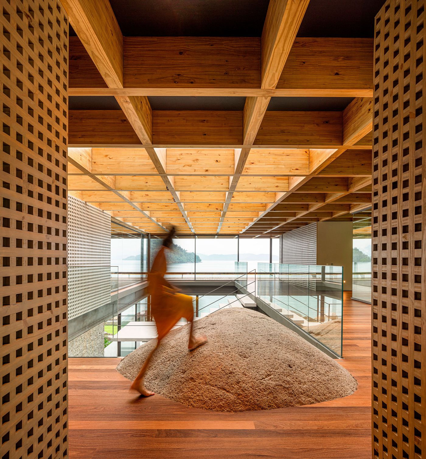 A dynamic indoor space features a remarkable natural boulder seamlessly integrated into the multi-level wooden flooring. A person in motion, dressed in yellow, ascends the stairs, highlighting the flow and connection between architectural elements and raw nature, all under an exposed timber ceiling grid and overlooking distant ocean views.
