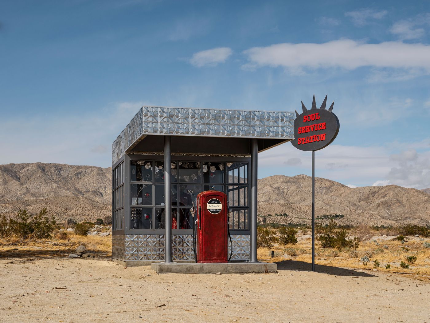 A unique red gas pump at a "Soul Service Station" in the desert, framed by mountains and a blue sky with scattered clouds.