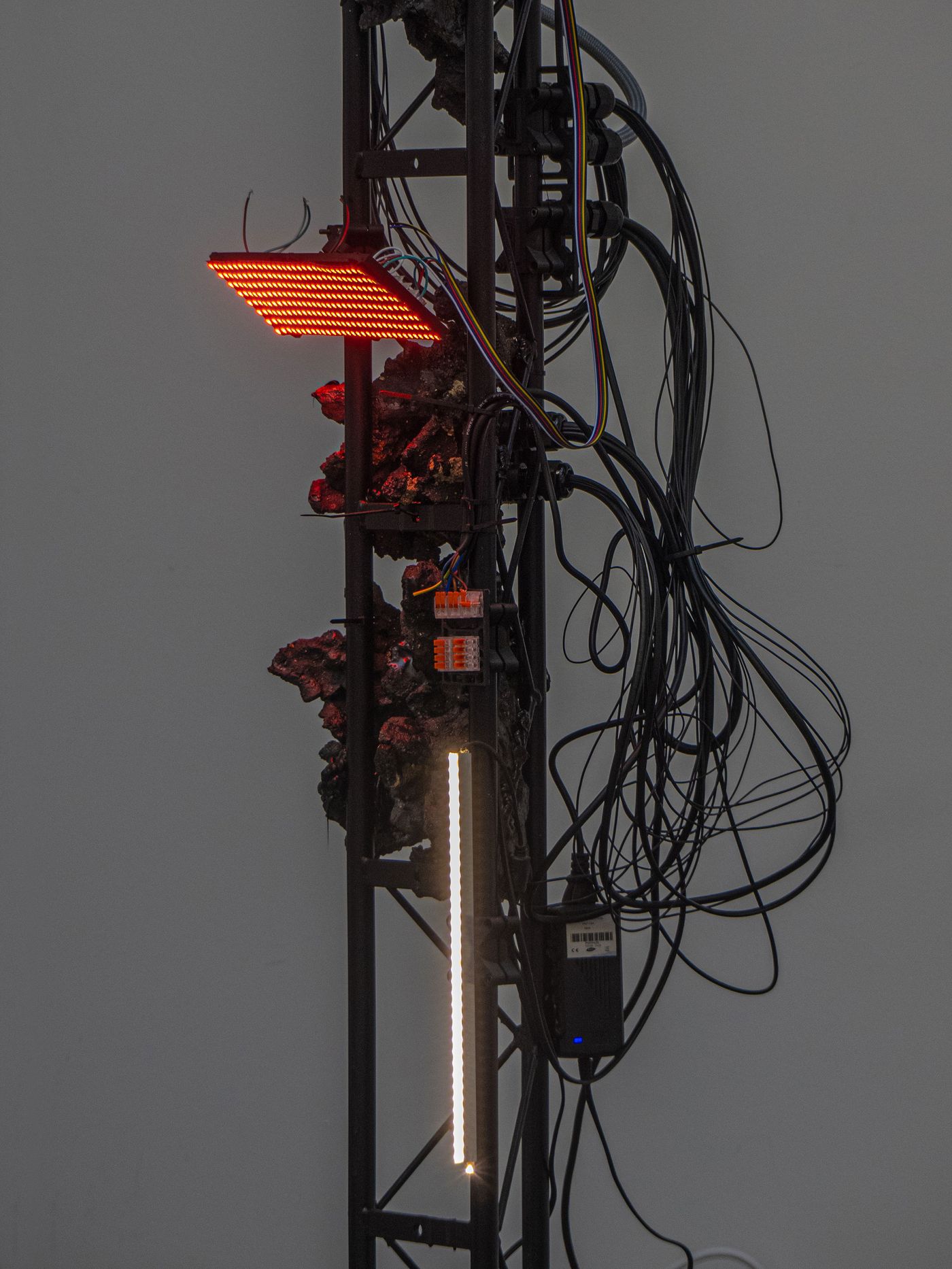 A vertical structure with red and white LED lights, entangled wires, and a textured surface, set against a plain wall.