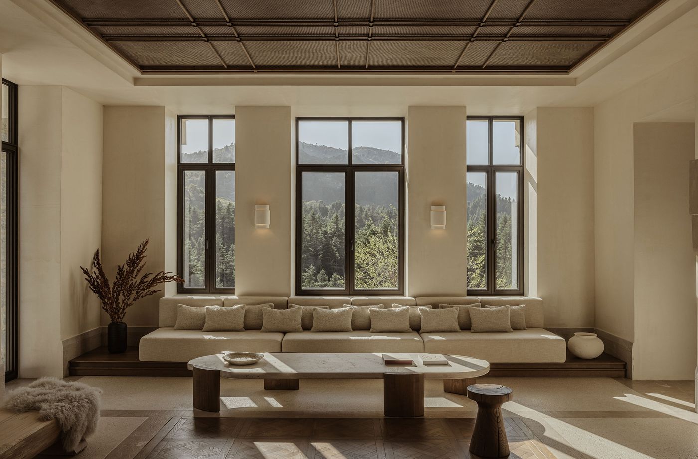 A spacious and serene common area, characterized by a long, low-slung built-in sofa with neutral-toned cushions, facing three tall, dark-framed windows that offer expansive views of a verdant forest. The room features a patterned ceiling, a contemporary oval coffee table, and warm natural light, embodying a sophisticated yet earthy aesthetic. A spacious and serene common area, characterized by a long, low-slung built-in sofa with neutral-toned cushions, facing three tall, dark-framed windows that offer expansive views of a verdant forest. The room features a patterned ceiling, a contemporary oval coffee table, and warm natural light, embodying a sophisticated yet earthy aesthetic.