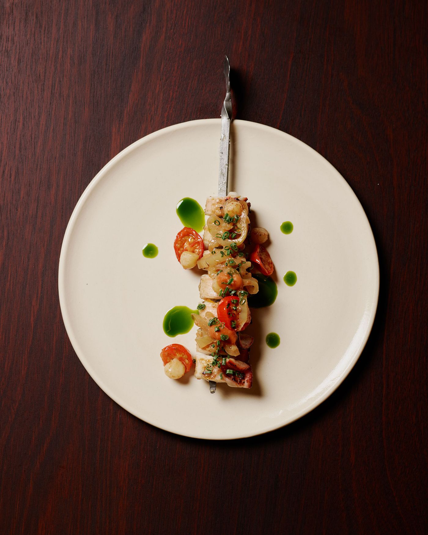A skewer of grilled seafood with scallops, shrimp, and cherry tomatoes drizzled with green sauce, served on a pale plate.