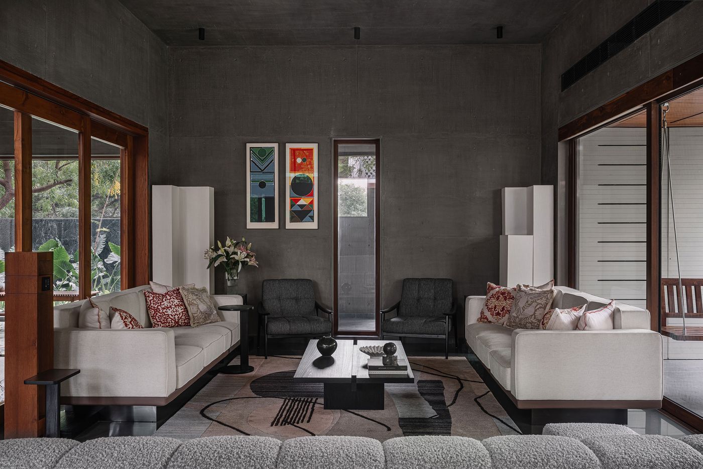 A grand living room defined by dark concrete walls and a polished floor, accented by a plush grey sectional sofa and two white sofas. A pair of grey armchairs adds a sophisticated touch. Abstract artworks with geometric shapes adorn the wall, while large windows connect the space to outdoor greenery, creating a harmonious blend of modern art and natural elements.