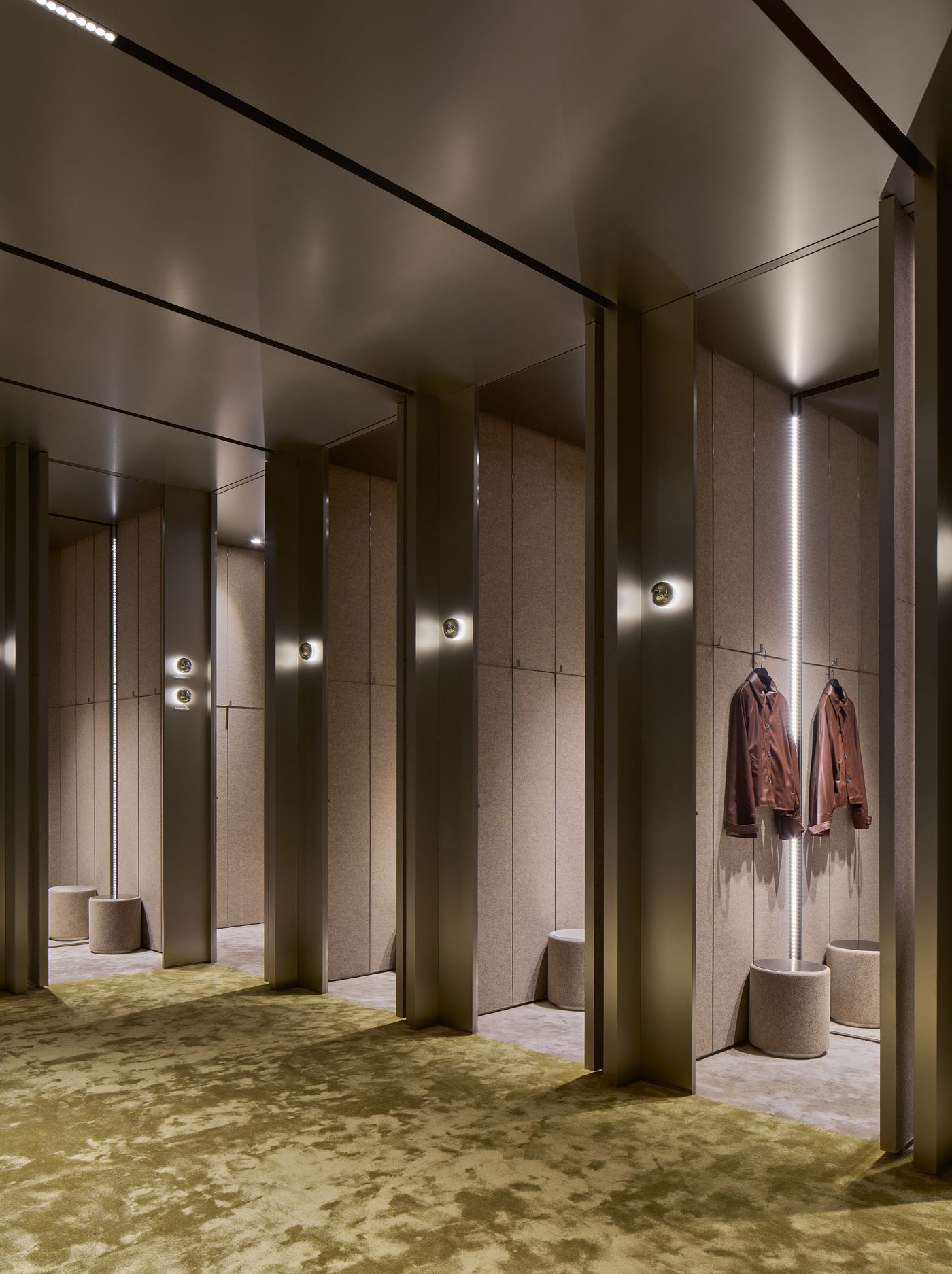 Modern dressing room featuring soft lighting, textured walls, and hanging coats, with plush green carpeting and circular seating.