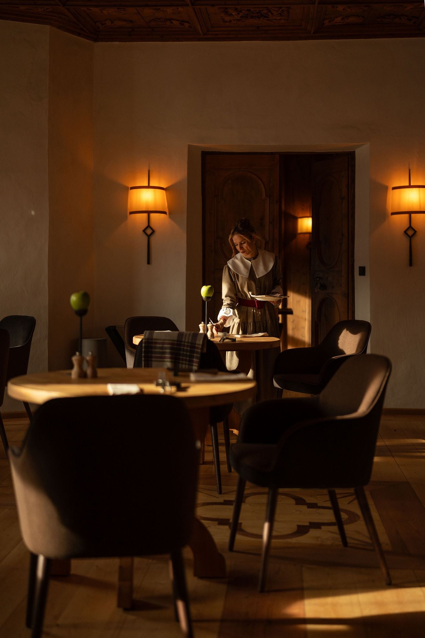 In a Castel Badia dining room, a server in a wool dress with a broad white collar and red belt sets a table in raking afternoon light. Parchment-shaded wall sconces on wrought-iron stems flank carved double doors, while green apples on slim stands and an inlaid parquet motif punctuate the room. In a Castel Badia dining room, a server in a wool dress with a broad white collar and red belt sets a table in raking afternoon light. Parchment-shaded wall sconces on wrought-iron stems flank carved double doors, while green apples on slim stands and an inlaid parquet motif punctuate the room.