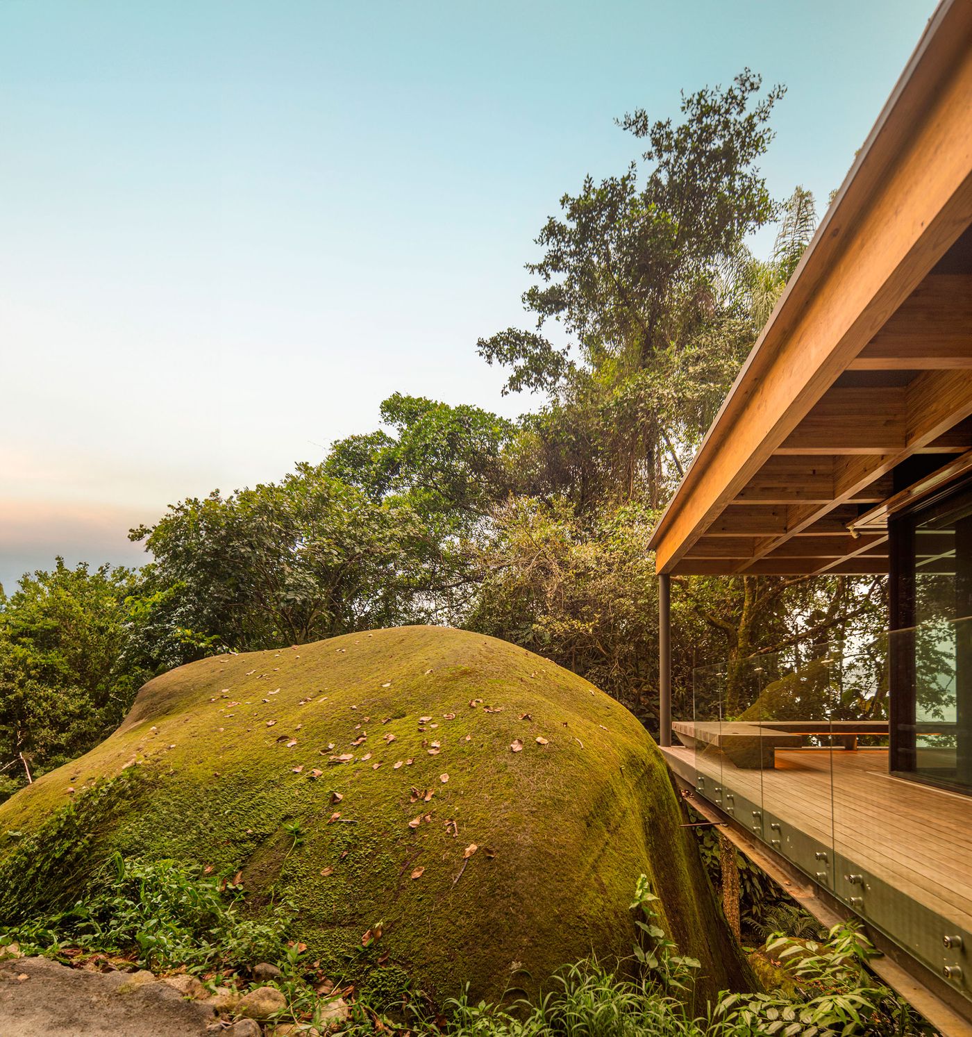 A grand moss-covered boulder dominates the foreground, juxtaposed with the elegant, low-slung residence and its expansive wooden deck with glass railings. The seamless connection between raw nature and refined architecture is highlighted by the surrounding lush tropical forest, under a clear, bright sky.