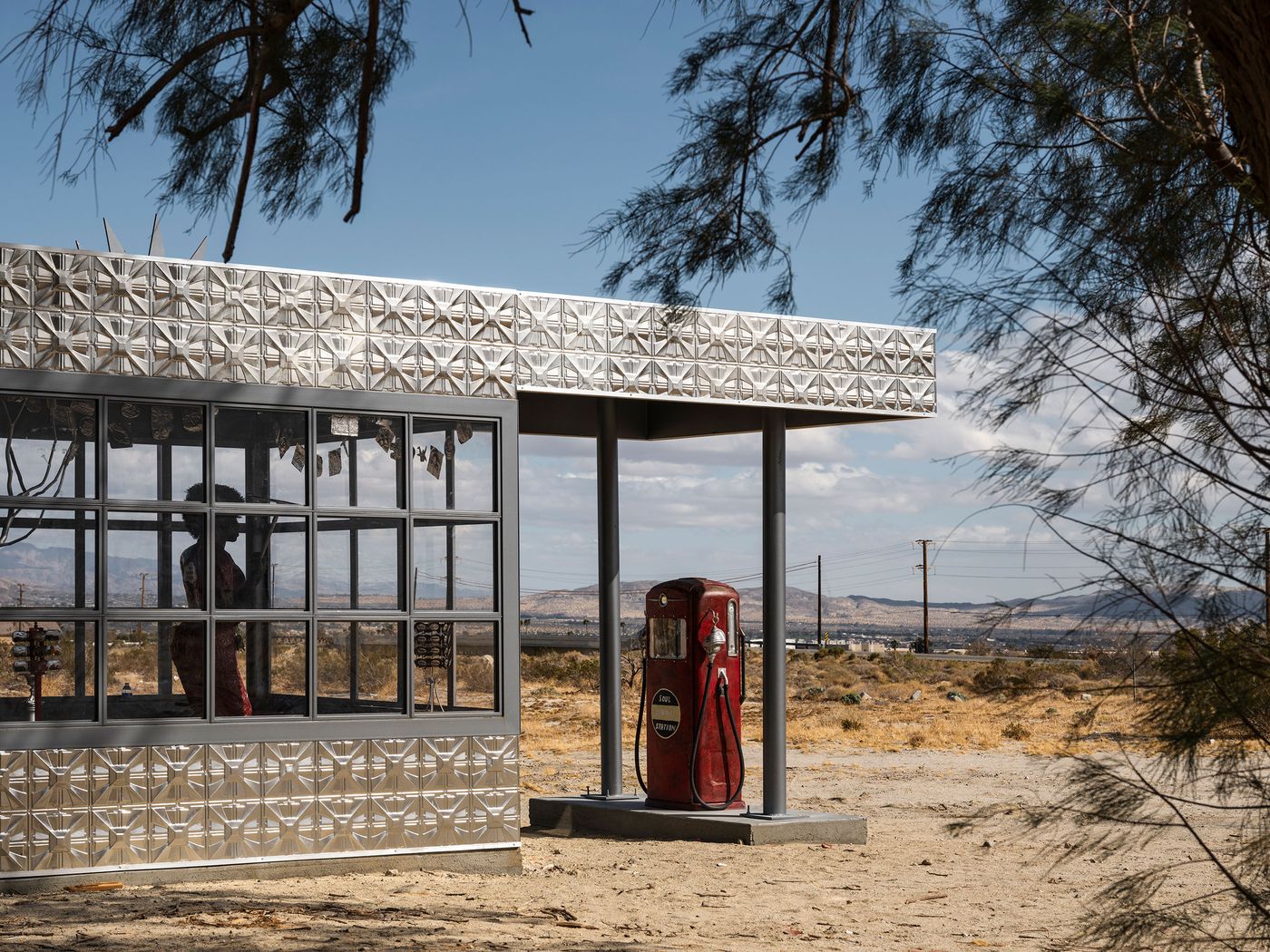 A vintage red gas pump stands beside a silver, modernist gas station in a desert landscape under a partly cloudy sky. A vintage red gas pump stands beside a silver, modernist gas station in a desert landscape under a partly cloudy sky.