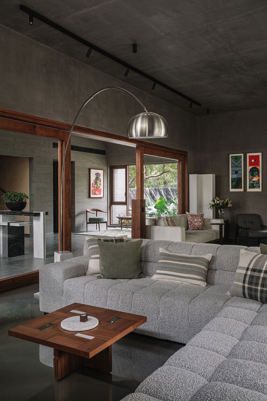 A luxurious living space features a large, textured grey sectional sofa adorned with patterned pillows. A striking arc floor lamp with a metallic dome shade illuminates the room, which boasts dark concrete walls and a dark floor. Warm wooden door frames lead to other areas, and abstract art pieces add vibrant pops of color, creating a refined and artistic ambiance.