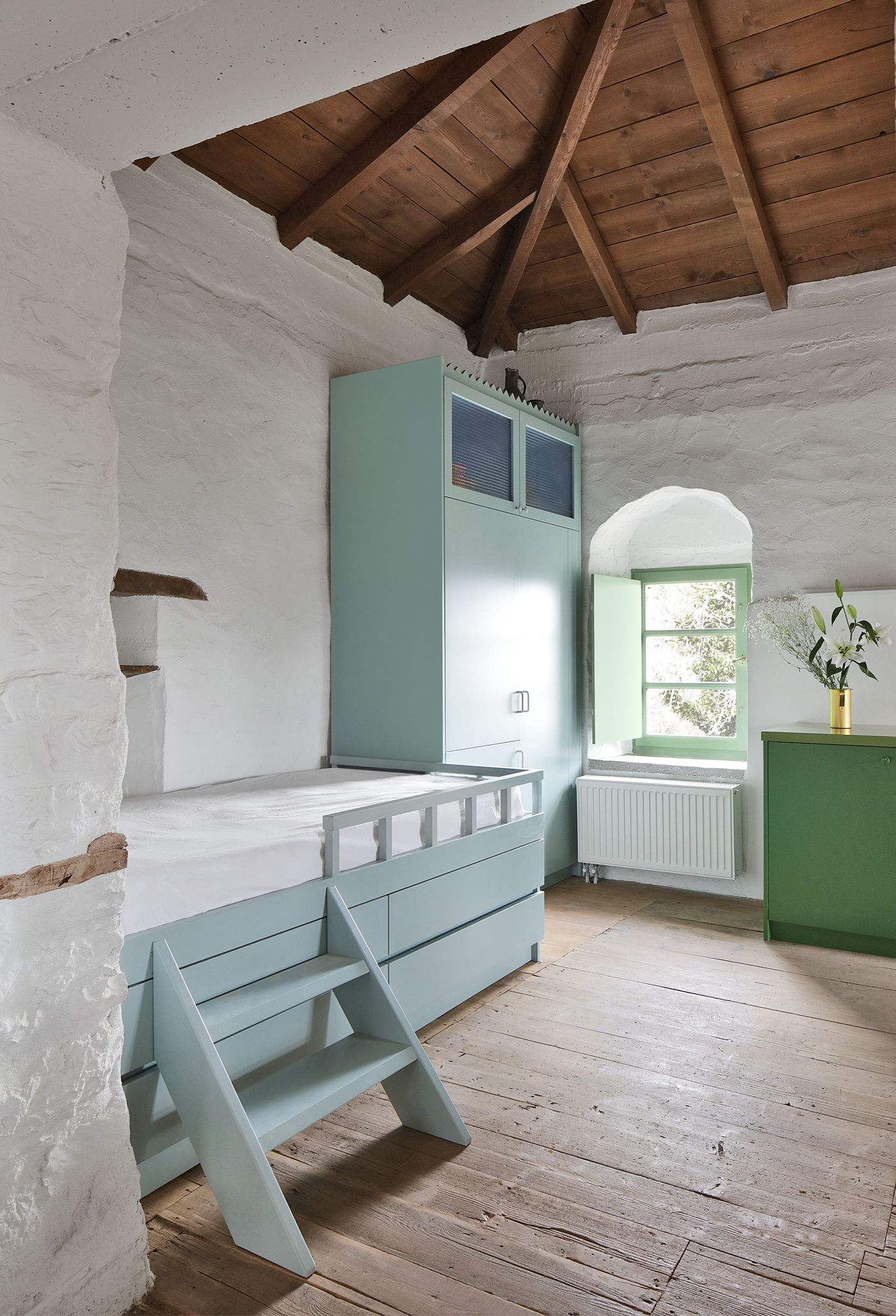 Sculptural sage-green staircase creates dynamic vertical circulation beneath the traditional timber roof, where built-in storage maximizes functionality within the compact space. The geometric contemporary intervention demonstrates how bold modern elements can enhance rather than compromise the building's authentic Mediterranean character and rustic material palette.