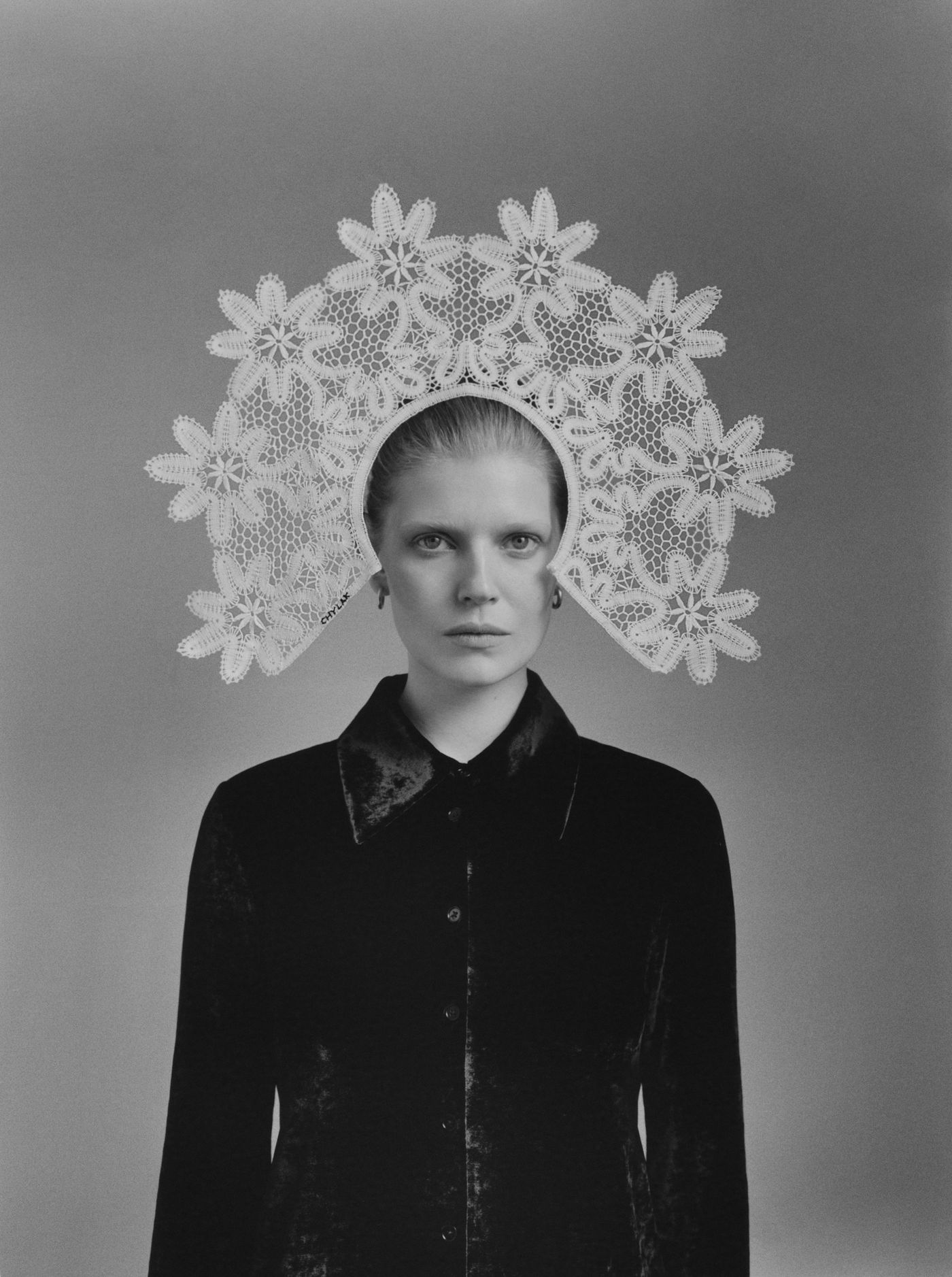 A figure wears a large white lace headdress, adorned with floral patterns, paired with a dark, stylish velvet shirt against a monochrome backdrop.
