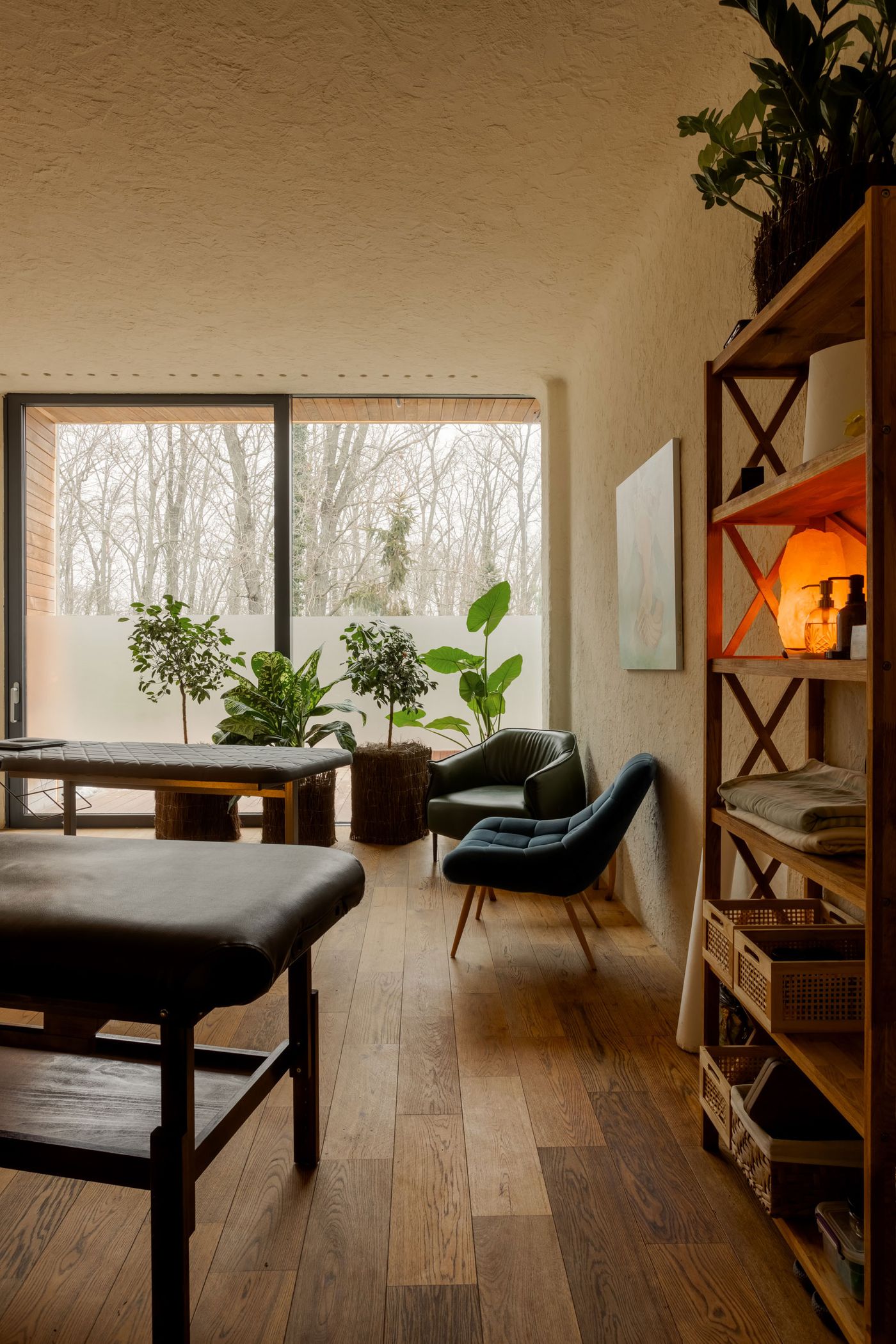 Cozy, modern interior featuring a massage table, plants, a green chair, wooden floor, and large windows with a nature view.