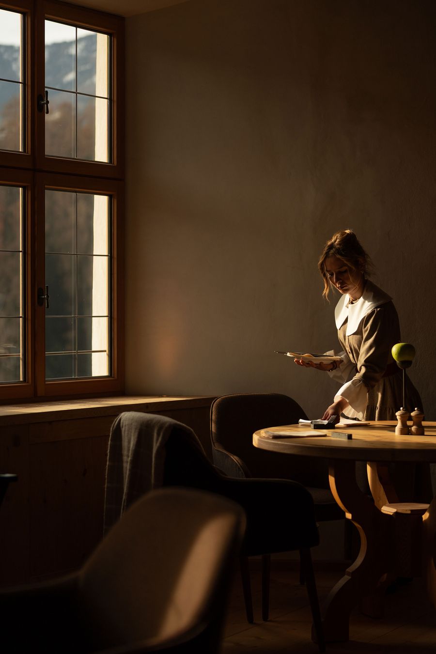 Low sun from a tall timber casement window catches a server laying cutlery on a round pine table at Castel Badia. The pared-back composition, with its plastered wall, sculpted pedestal base, wooden pepper mills and a single green apple, has the quiet, painterly stillness of a Dutch interior. Low sun from a tall timber casement window catches a server laying cutlery on a round pine table at Castel Badia. The pared-back composition, with its plastered wall, sculpted pedestal base, wooden pepper mills and a single green apple, has the quiet, painterly stillness of a Dutch interior.