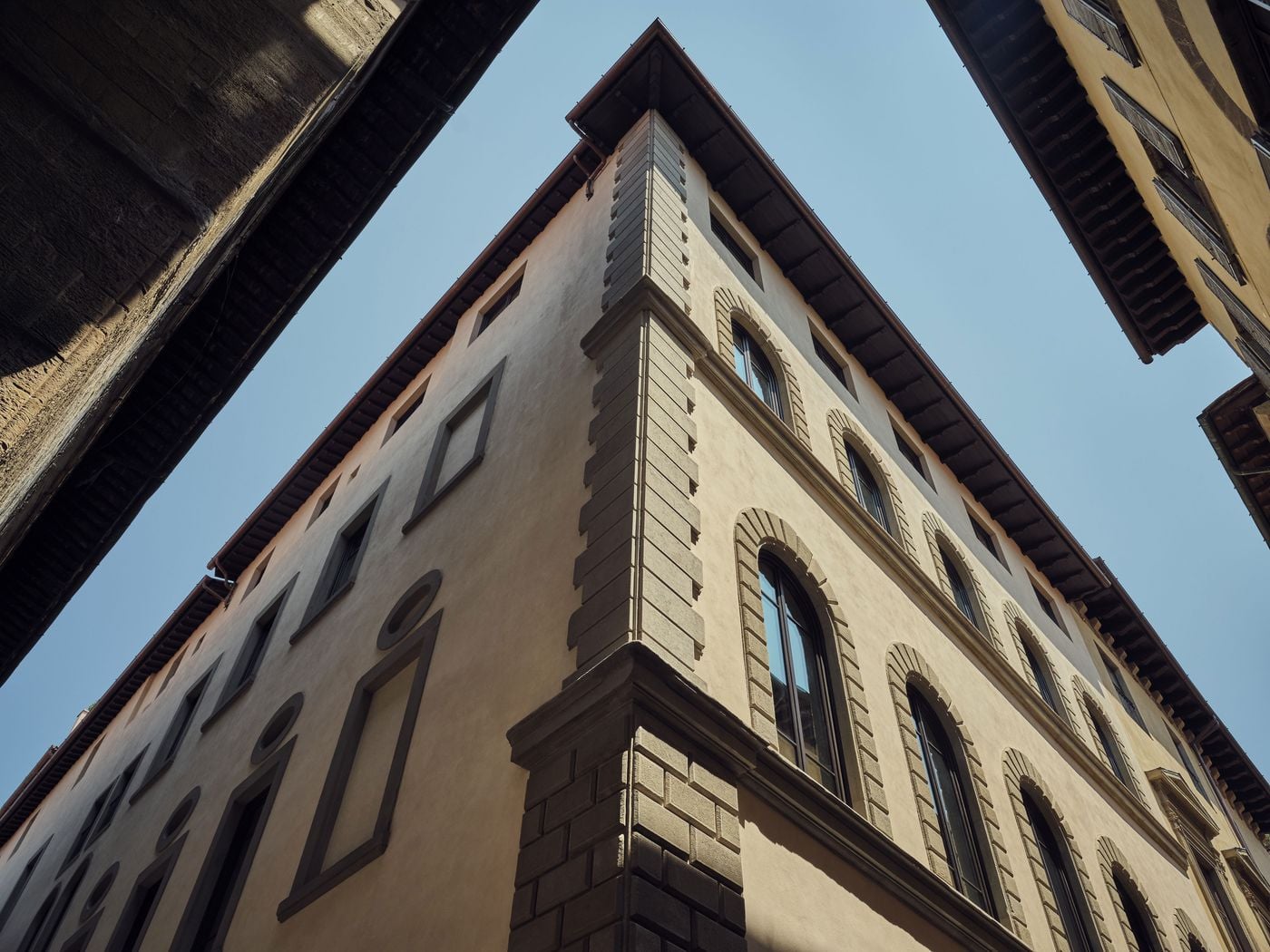 Looking up at the corner of La Réserve Firenze, rusticated quoins climb towards deep timber eaves, framed against a strip of blue sky by the overhanging roofs of neighbouring buildings. Arched windows, oculi and stone-framed blind panels animate the warm ochre facade. Looking up at the corner of La Réserve Firenze, rusticated quoins climb towards deep timber eaves, framed against a strip of blue sky by the overhanging roofs of neighbouring buildings. Arched windows, oculi and stone-framed blind panels animate the warm ochre facade.
