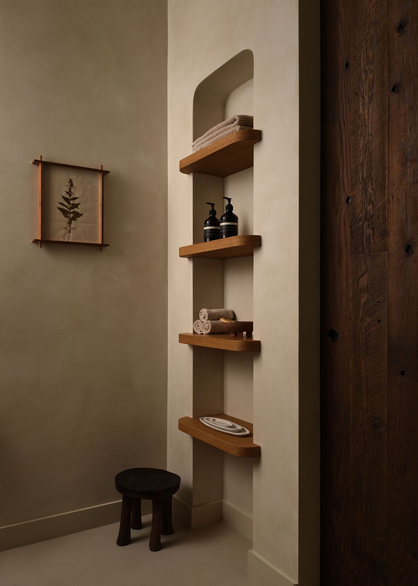 Minimalist bathroom corner featuring wooden shelves, towels, dark soap dispensers, a small stool, and a framed botanical print. Minimalist bathroom corner featuring wooden shelves, towels, dark soap dispensers, a small stool, and a framed botanical print.