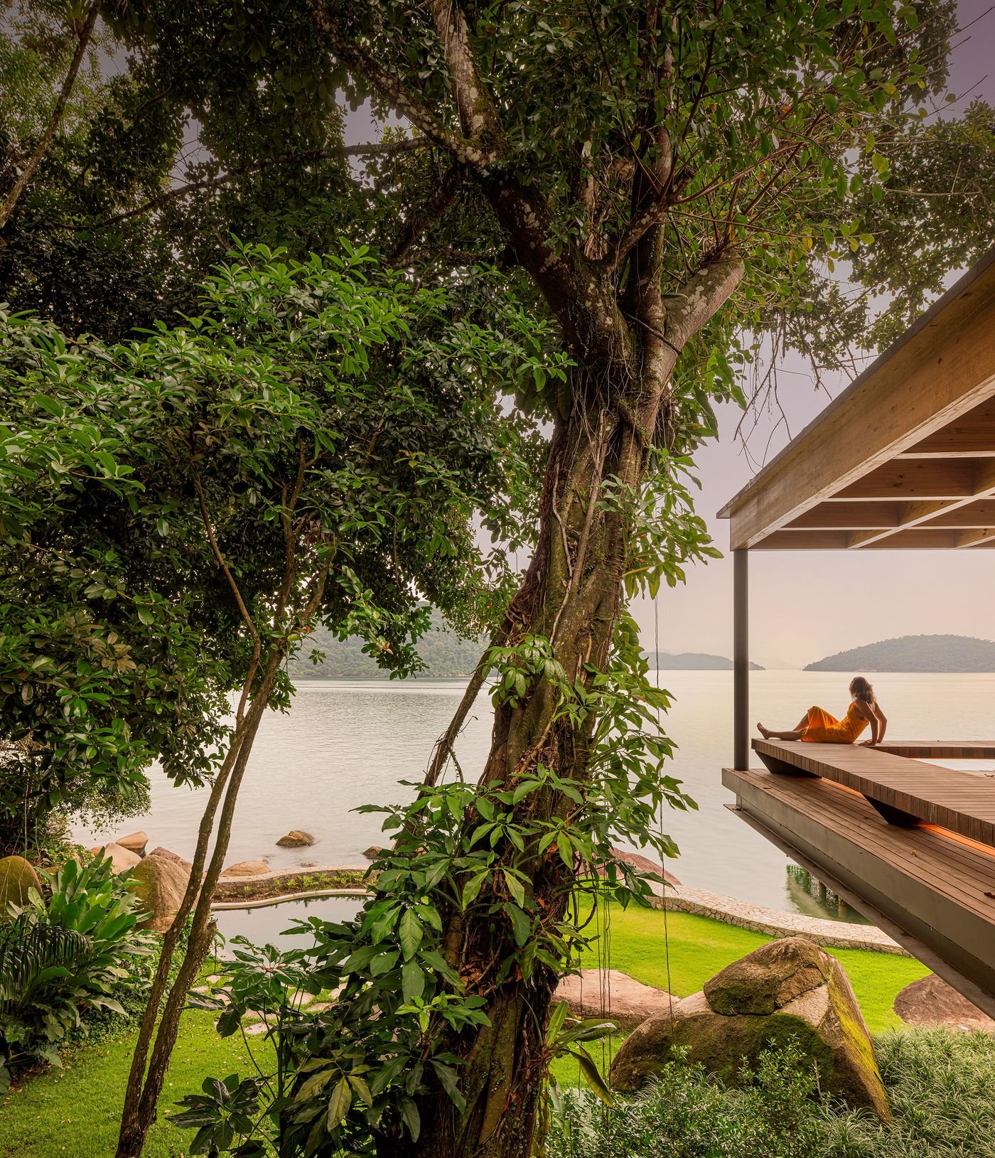 A person in a yellow dress relaxes on a cantilevered wooden deck, overlooking serene waters framed by lush tropical trees and natural rock formations. The design harmonizes with the coastal landscape, providing a tranquil outdoor living space.