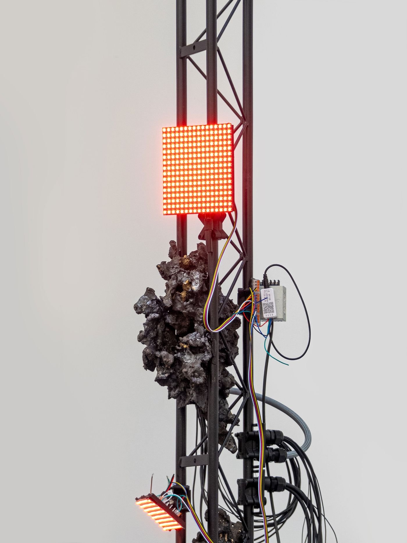 A close-up of a metallic structure featuring red LED lights and tangled wires, alongside a textured, rocky material. A close-up of a metallic structure featuring red LED lights and tangled wires, alongside a textured, rocky material.