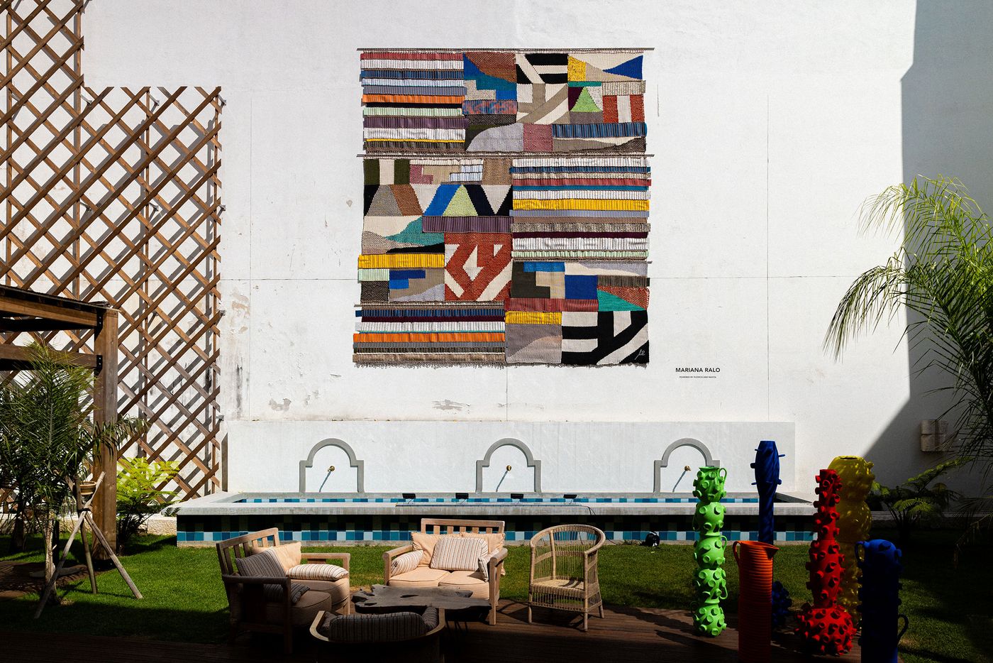 An outdoor courtyard at Locke Santa Joana features a striking, large-scale textile artwork by Mariana Ralo, composed of abstract, colorful geometric patterns. Below, a classic white fountain with three spouts anchors a lush green lawn, complemented by natural material seating. Tall, vibrant, sculptural forms by Terrakota studio add a modern, eclectic touch to this serene outdoor setting. An outdoor courtyard at Locke Santa Joana features a striking, large-scale textile artwork by Mariana Ralo, composed of abstract, colorful geometric patterns. Below, a classic white fountain with three spouts anchors a lush green lawn, complemented by natural material seating. Tall, vibrant, sculptural forms by Terrakota studio add a modern, eclectic touch to this serene outdoor setting.