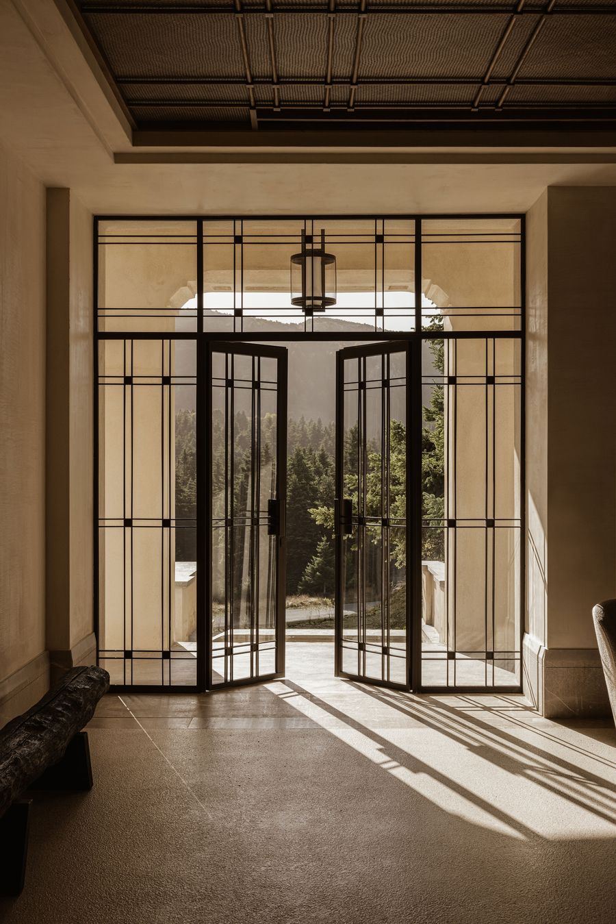 A grand entrance features tall, dark-framed glass doors and windows, creating a striking visual grid. The doors are open, revealing a lush, green forest beyond, while sunbeams cast dramatic shadows across the light-colored terrazzo floor. A minimalist hanging lantern provides a touch of classic elegance within this modern design. A grand entrance features tall, dark-framed glass doors and windows, creating a striking visual grid. The doors are open, revealing a lush, green forest beyond, while sunbeams cast dramatic shadows across the light-colored terrazzo floor. A minimalist hanging lantern provides a touch of classic elegance within this modern design.