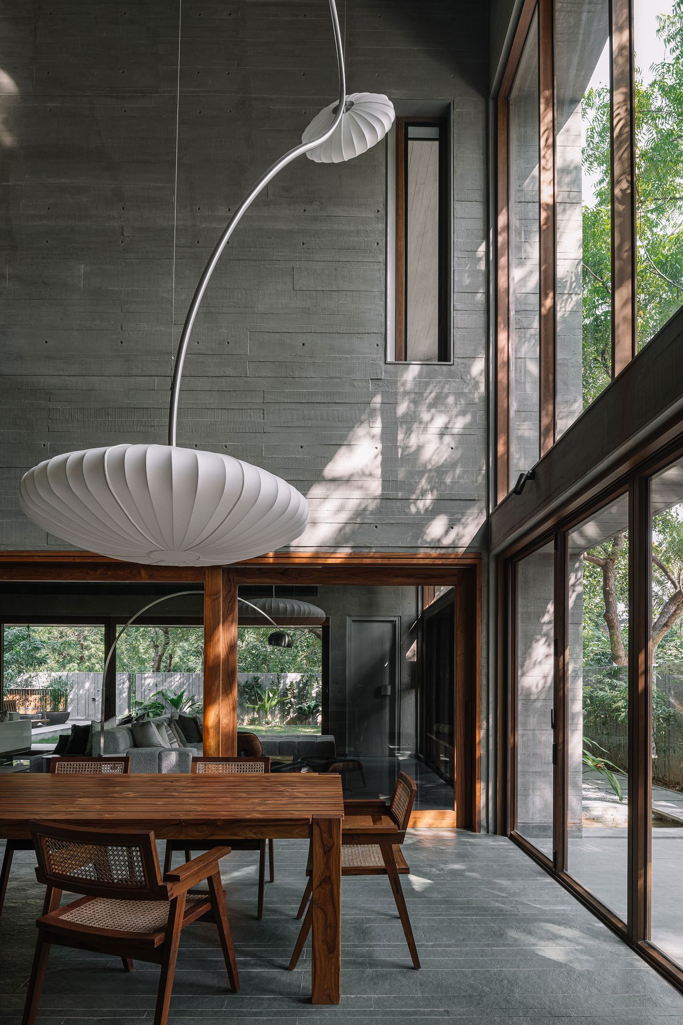 A captivating dining area bathed in natural light, featuring a large, elegant wooden dining table and rattan chairs. A sculptural, pleated white pendant light floats above, adding a touch of softness to the raw concrete walls and expansive, wood-framed windows that seamlessly blend the interior with the lush outdoor greenery.