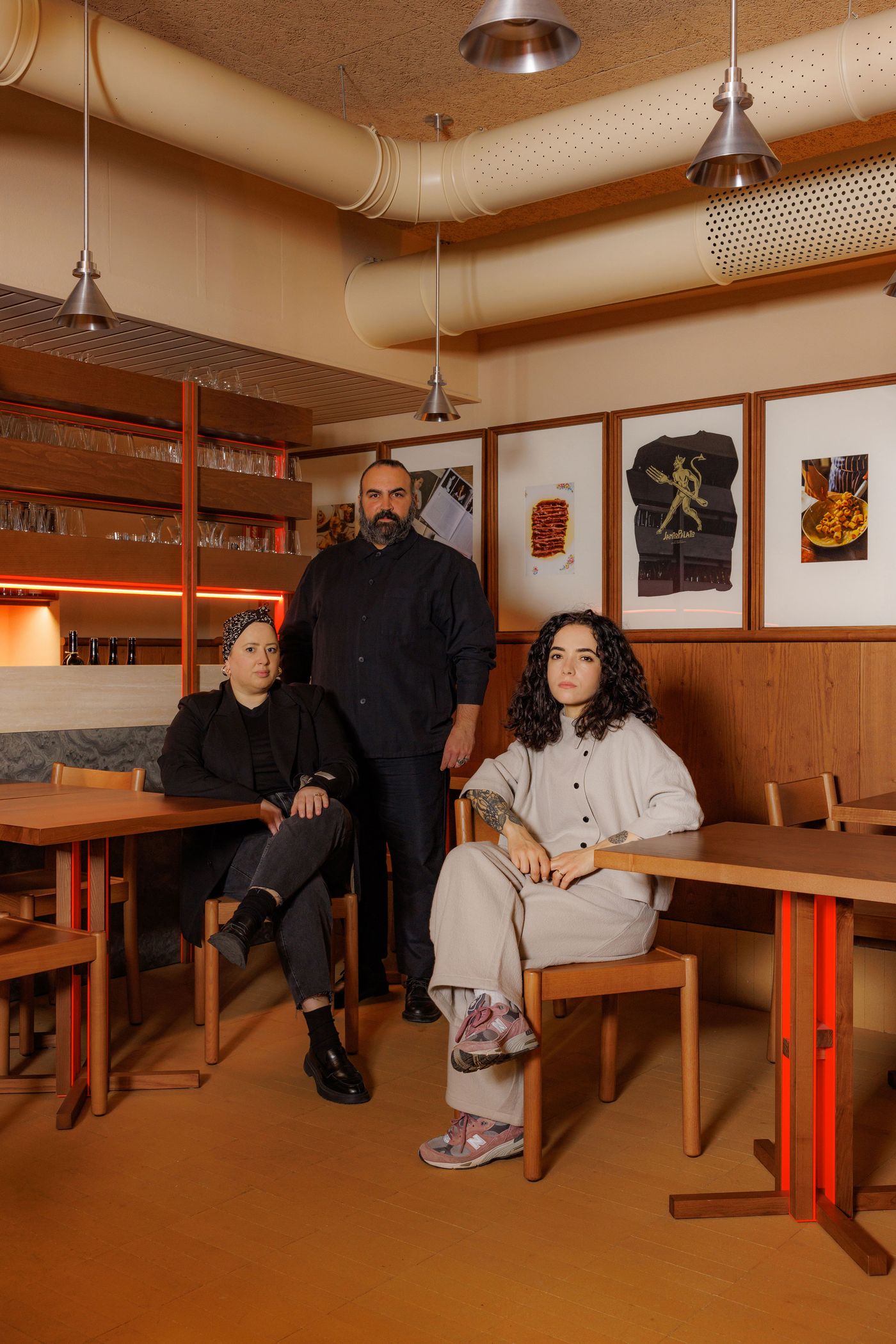 A man and two women pose amidst a meticulously crafted restaurant space. Surrounded by warm wood paneling, framed food art, and sleek tables with bold red bases, they embody the fusion of modern design and authentic Roman sensibility. The overhead industrial pendant lights illuminate the inviting, artistic atmosphere. A man and two women pose amidst a meticulously crafted restaurant space. Surrounded by warm wood paneling, framed food art, and sleek tables with bold red bases, they embody the fusion of modern design and authentic Roman sensibility. The overhead industrial pendant lights illuminate the inviting, artistic atmosphere.