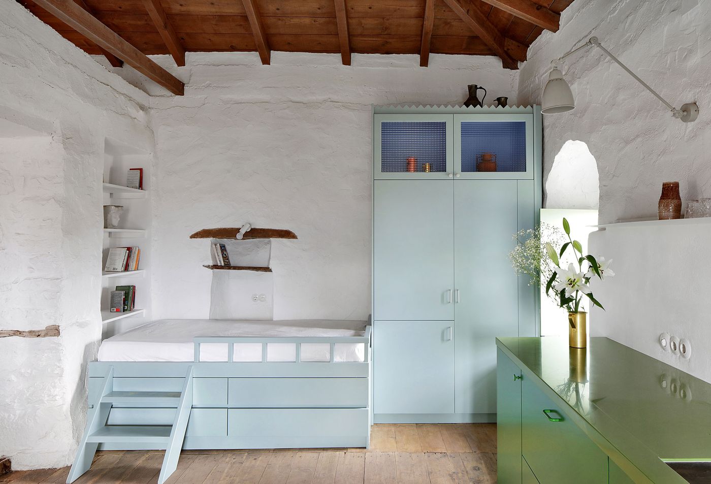 Modular sage-green furniture creates flexible living arrangements within the whitewashed stone interior, where perforated metal screens provide visual texture while maintaining storage functionality. The built-in shelving and contemporary sleeping platform demonstrate thoughtful space planning that respects the building's historical proportions and timber ceiling structure.