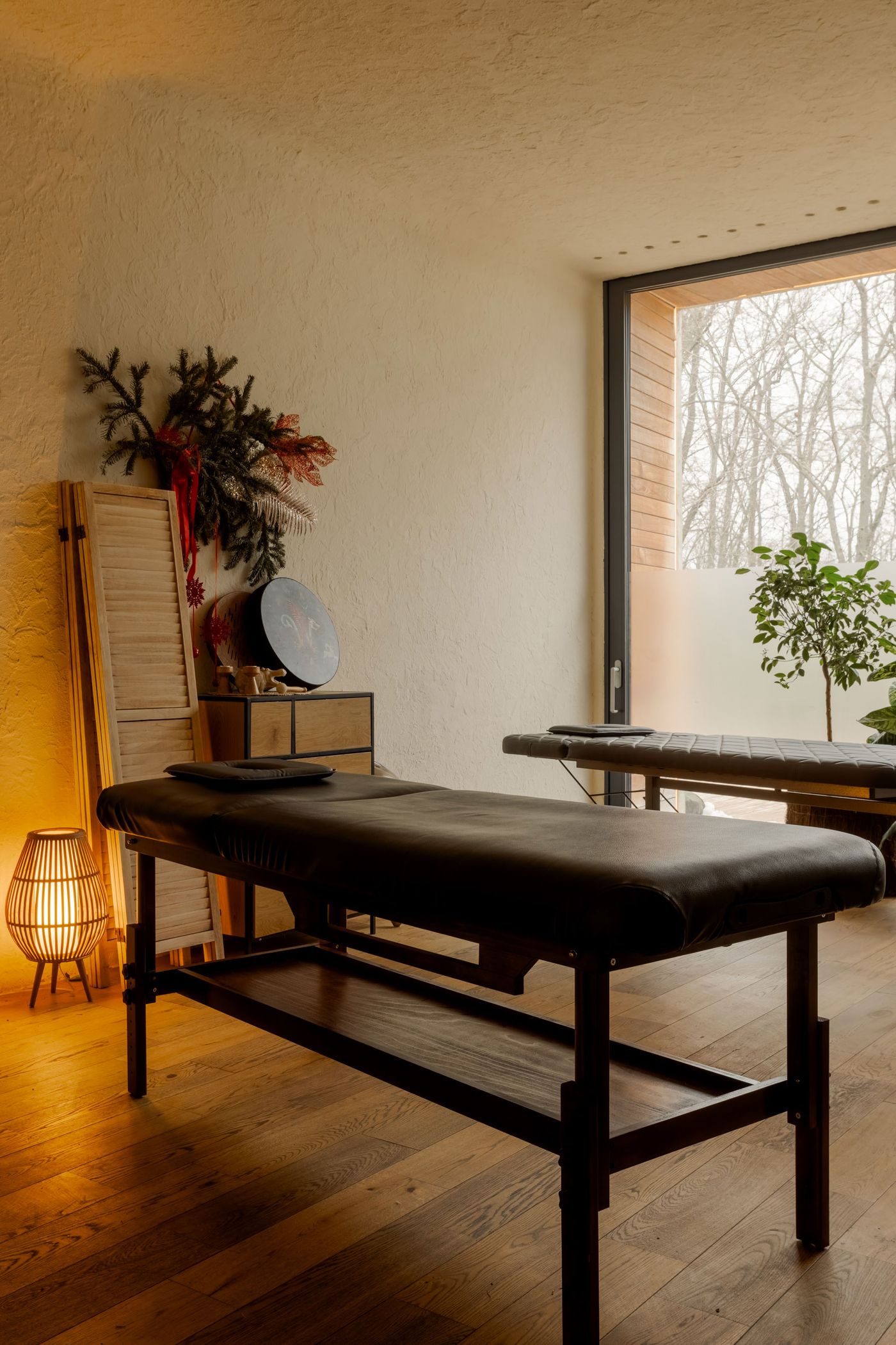 A serene massage therapy room featuring a black massage table, soft lighting, natural decor, and a window with a view of trees.